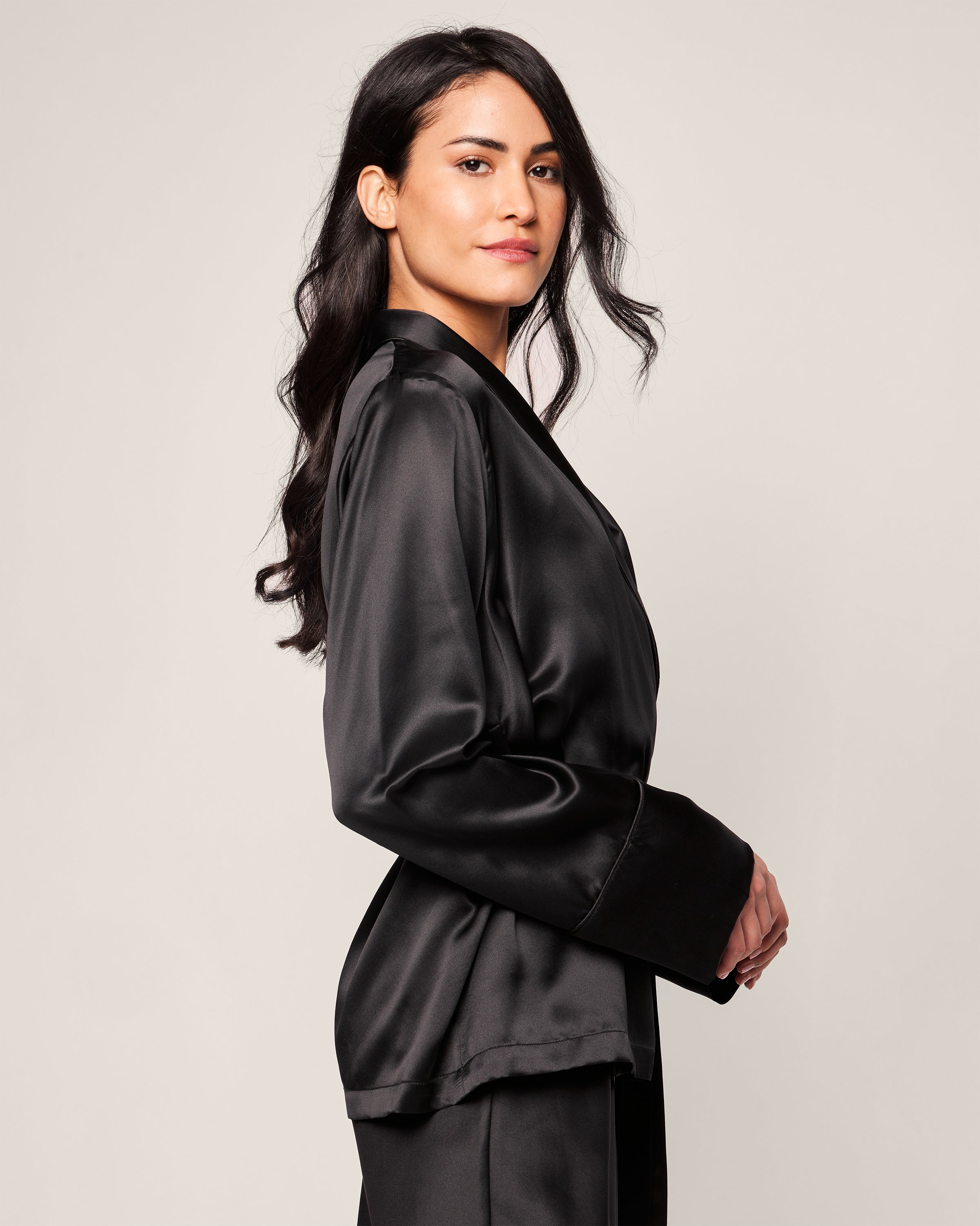 A person with long, dark hair stands sideways, glancing over their shoulder in a black Womens Silk Short Robe by Petite Plume, exuding elegance and hypoallergenic comfort. The plain, light background enhances their confident presence.