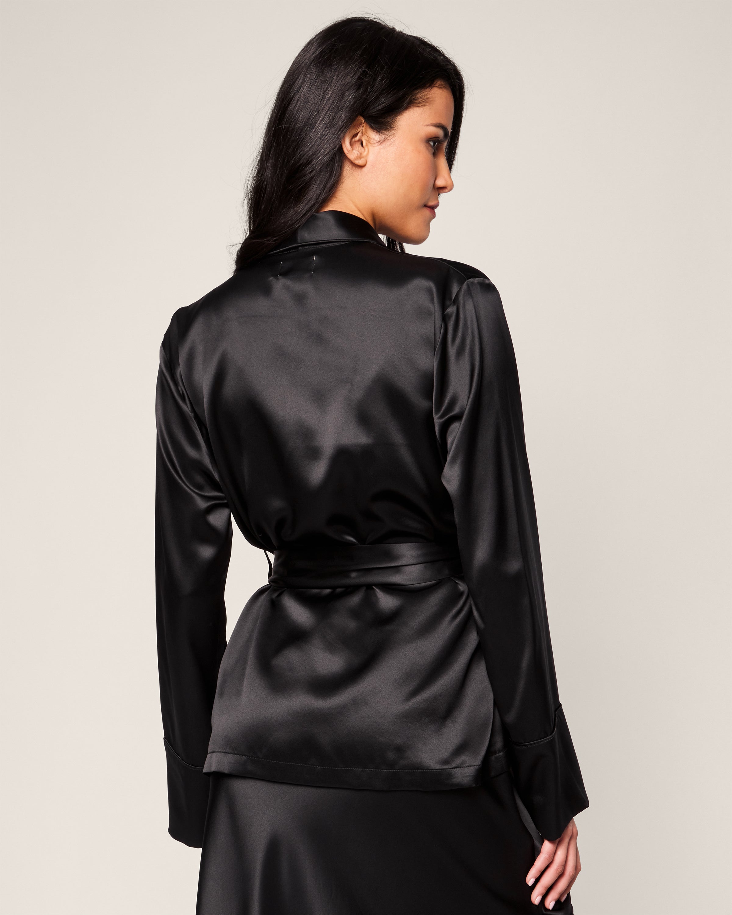 A woman with long dark hair faces away from the camera, wearing Petite Plumes Womens Silk Short Robe in Black, made from hypoallergenic Mulberry Silk. The plain light background highlights the elegance of her attire.