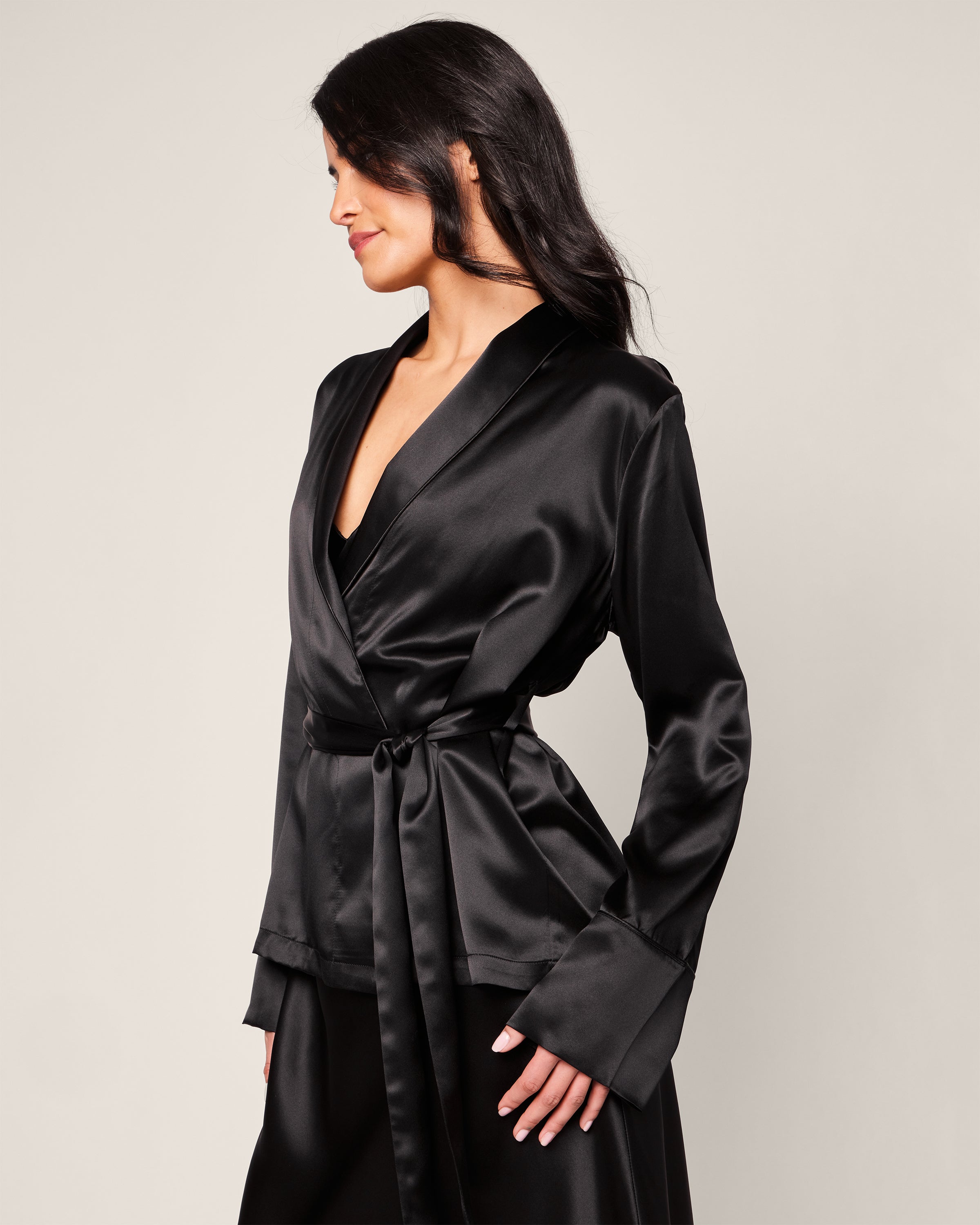A person with long, dark hair models the Womens Silk Short Robe in Black by Petite Plume, featuring a deep V-neck and tied waist. They stand sideways against a gray background, showcasing an air of luxurious sleepwear sophistication.