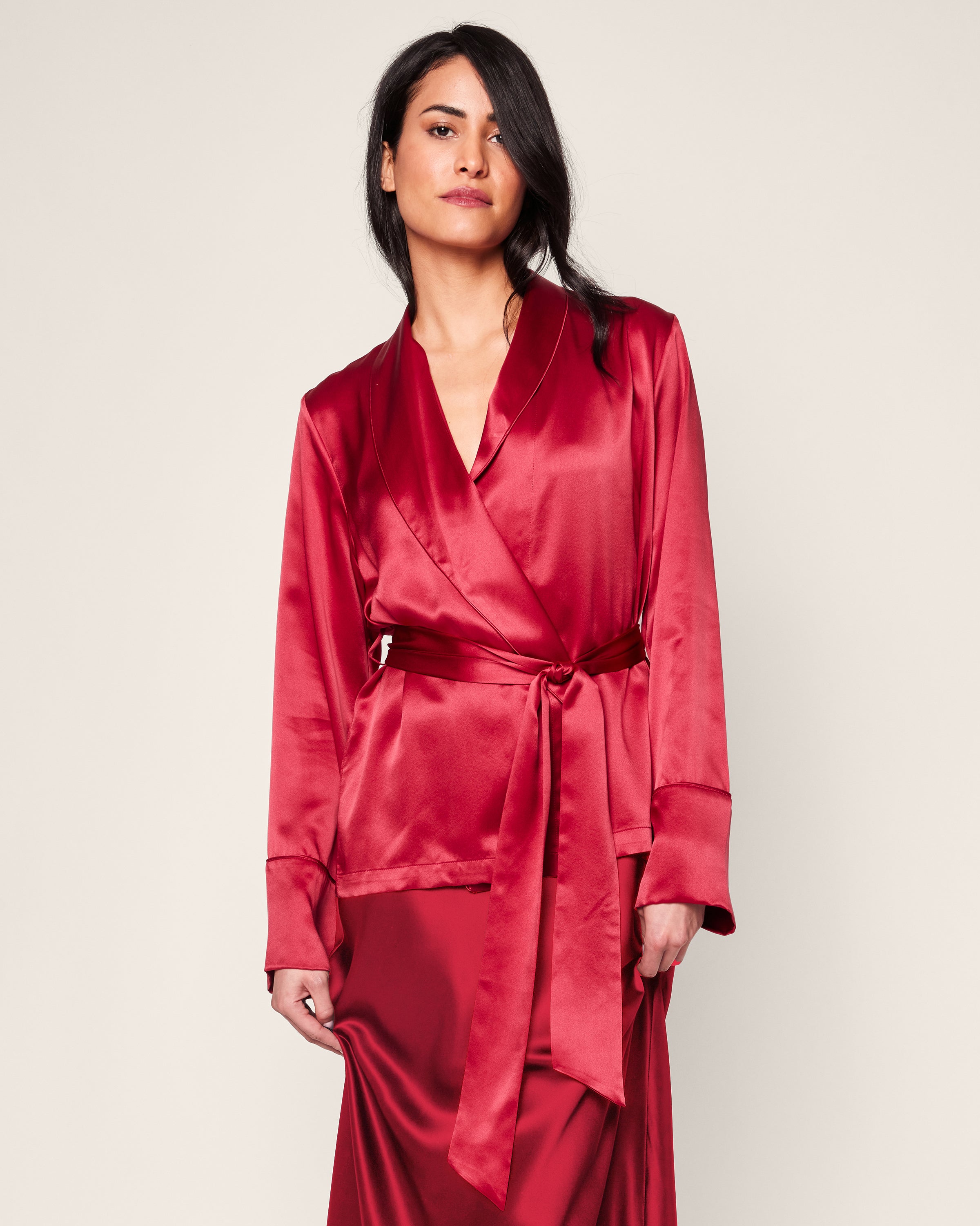 A person in a crimson Womens Silk Short Robe by Petite Plume stands against a neutral background, with long dark hair and a sidelong gaze.