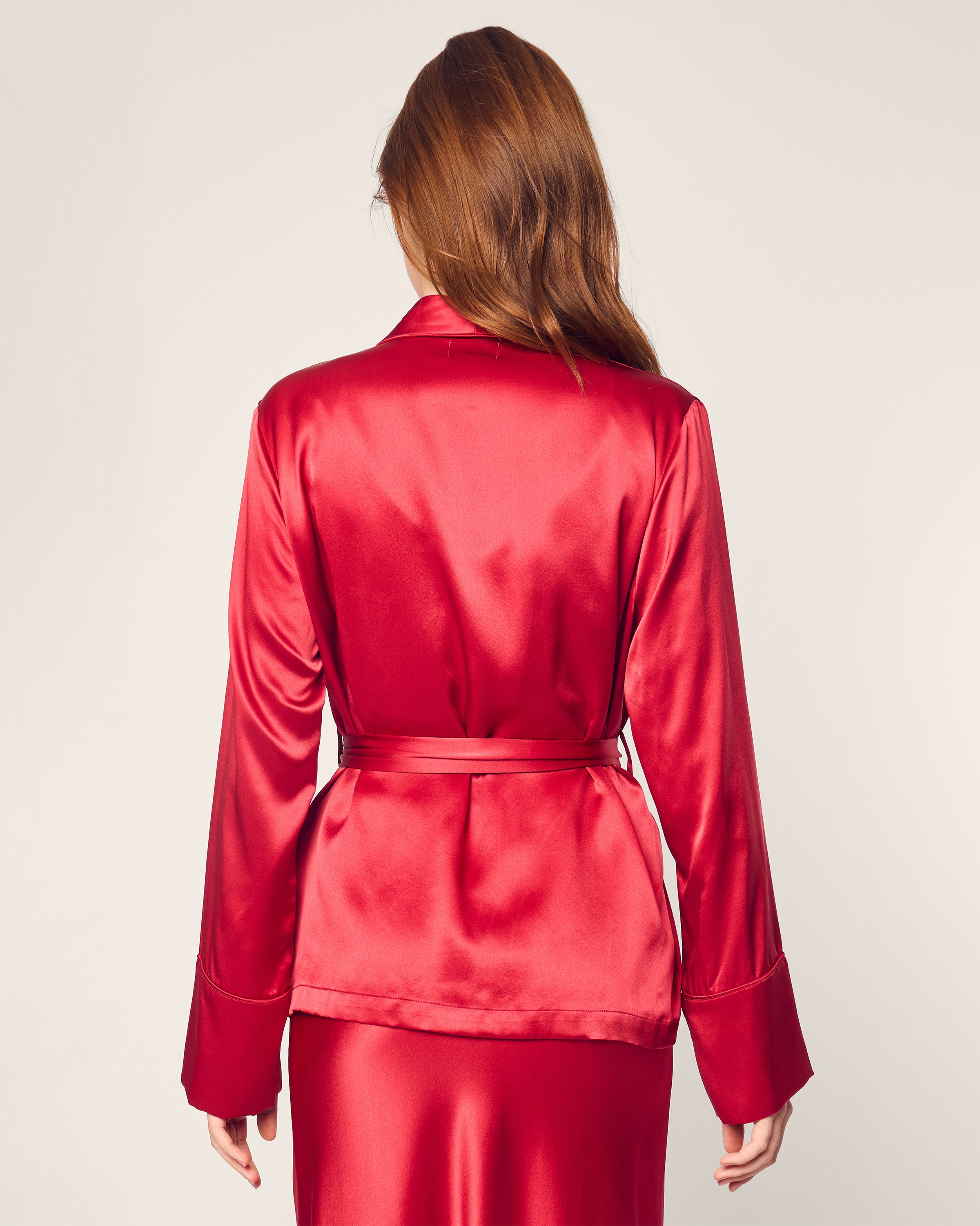 A person with long brown hair is shown from the back wearing the Womens Silk Short Robe in Crimson by Petite Plume. The shiny robe has long sleeves and a waist belt, set against a light, plain background that enhances its luxuriousness.
