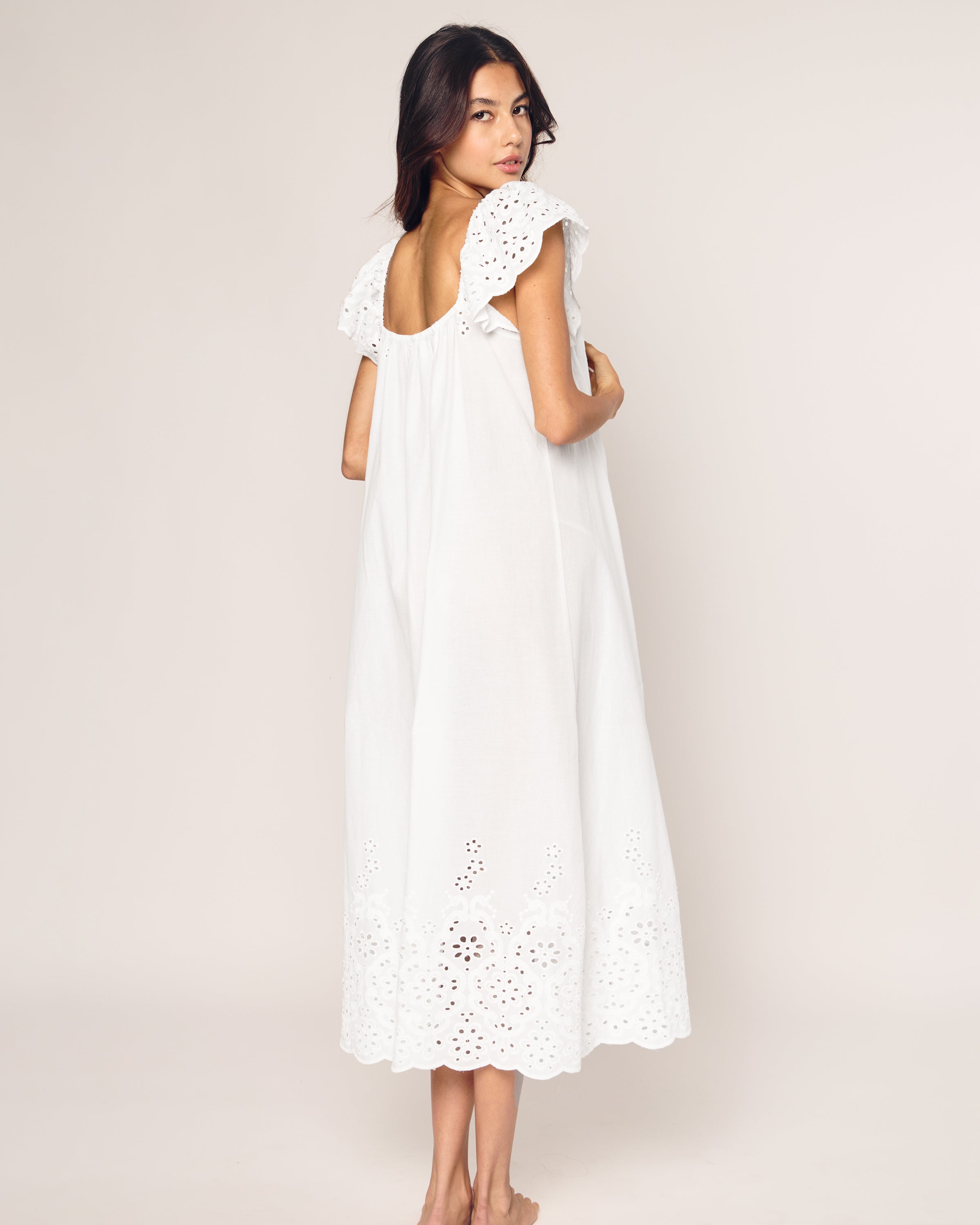A person stands barefoot, facing away, in a white sleeveless Womens Twill Ava Nightgown by Petite Plume, adorned with intricate eyelet embroidery on the hem and shoulder frills. Made from premium cotton, this elegant sleepwear beautifully contrasts with the plain off-white background.