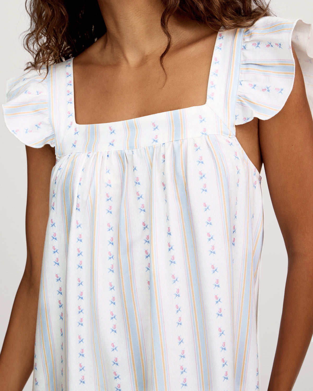 A woman stands barefoot against a plain light background wearing the Women's Twill Clara Nightgown in Cottage Stripe, featuring a long, loose fit with pastel vertical stripes and ruffled shoulder straps.