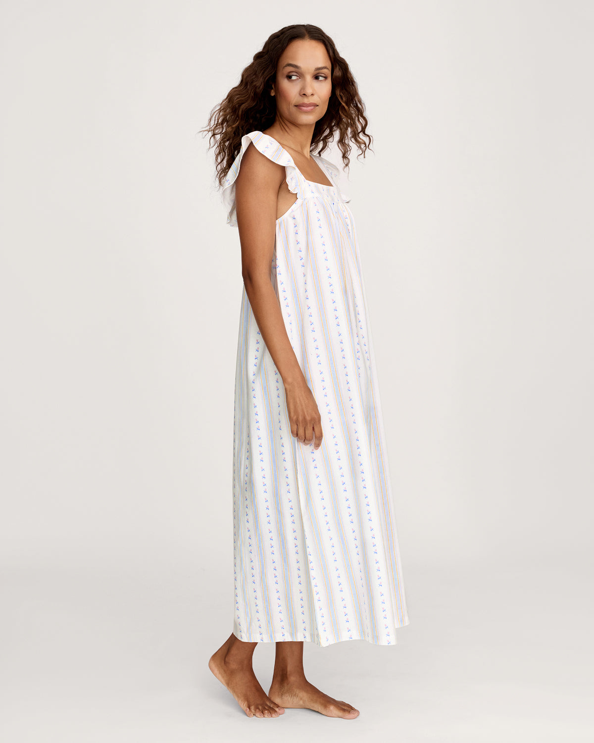 A woman stands barefoot against a plain light background wearing the Women's Twill Clara Nightgown in Cottage Stripe, featuring a long, loose fit with pastel vertical stripes and ruffled shoulder straps.