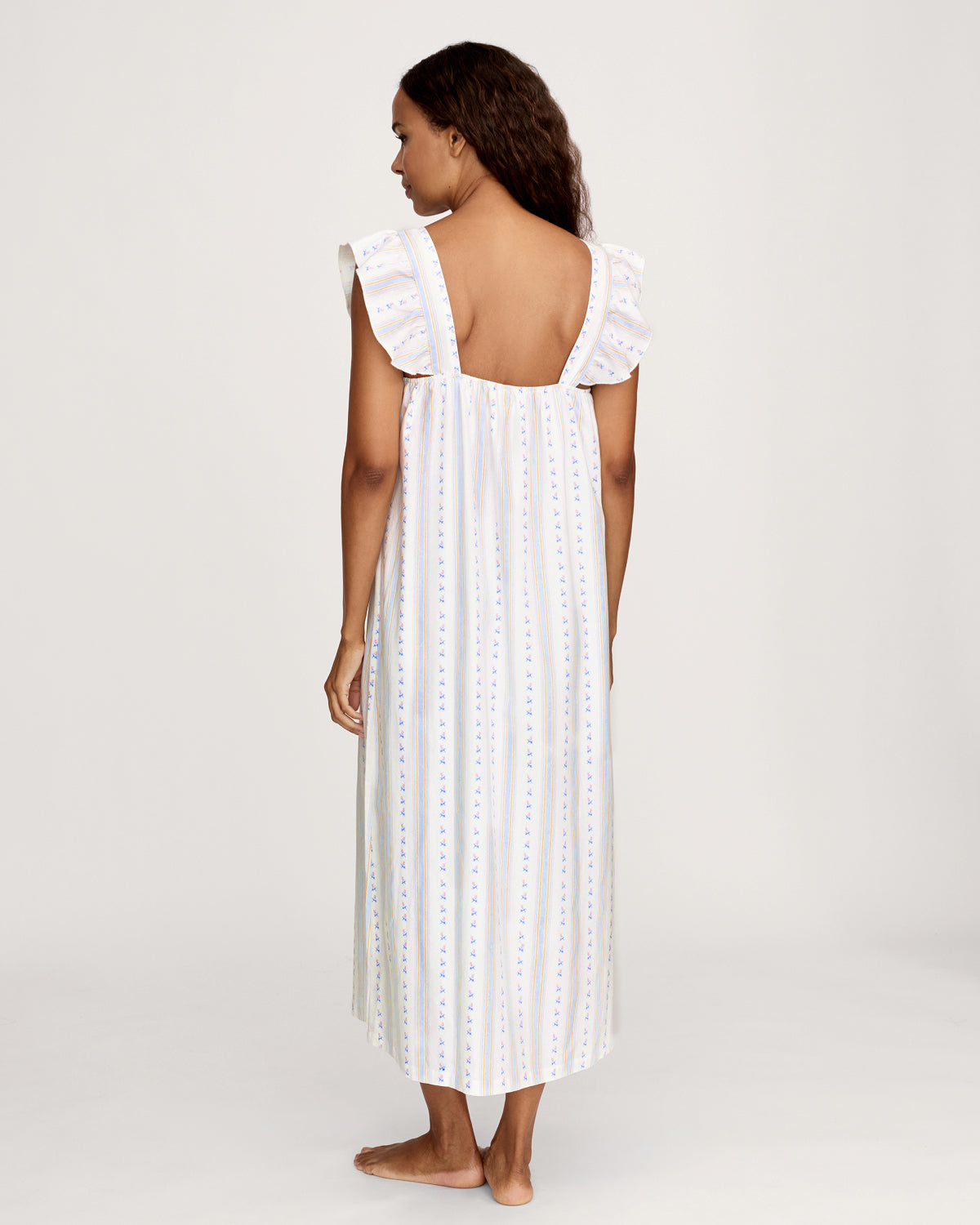 A woman stands barefoot against a plain light background wearing the Women's Twill Clara Nightgown in Cottage Stripe, featuring a long, loose fit with pastel vertical stripes and ruffled shoulder straps.