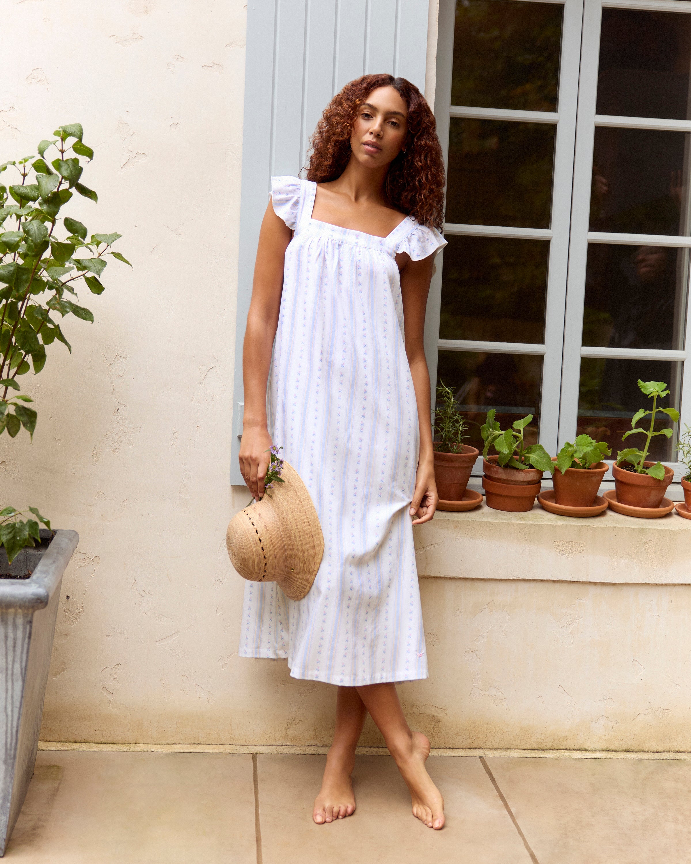 A woman stands barefoot against a plain light background wearing the Women's Twill Clara Nightgown in Cottage Stripe, featuring a long, loose fit with pastel vertical stripes and ruffled shoulder straps.