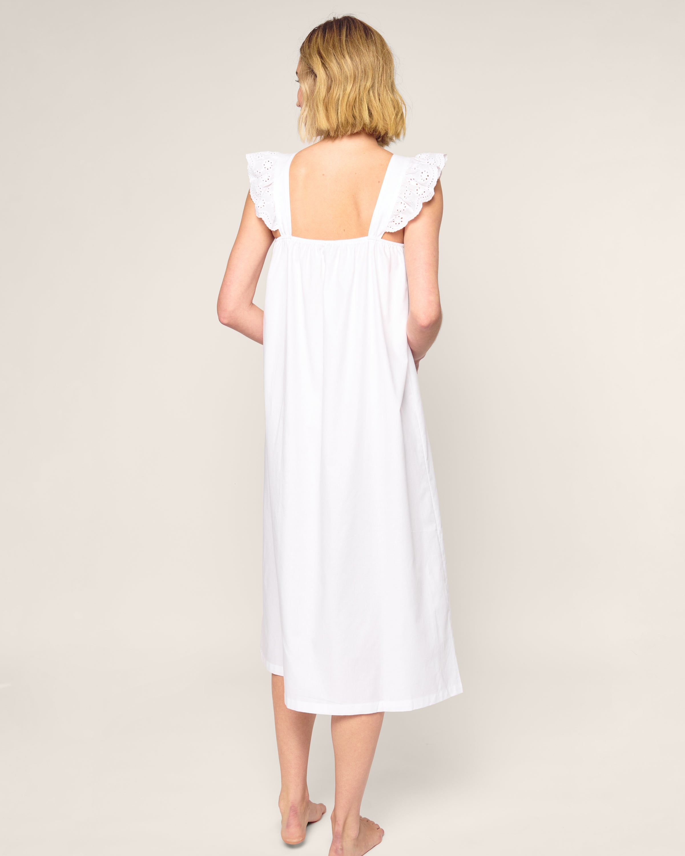 A person with short blonde hair stands barefoot on a neutral background, wearing the luxurious Womens Twill Clara Nightgown in White by Petite Plume. This sleeveless, knee-length dress has lace detailing on the shoulders and back, crafted from top-quality cotton. The back is visible.