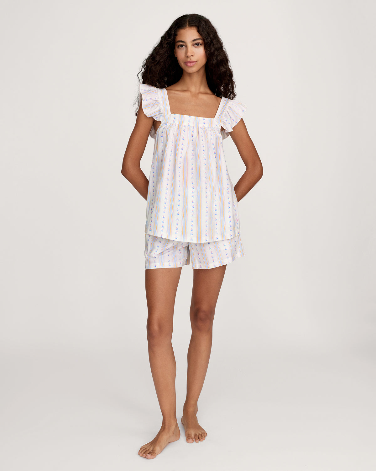 A woman with long curly hair models the Women's Twill Clara Short Set in Cottage Stripe—a loose, sleeveless pajama set with ruffled shoulders and shorts, featuring white fabric with pastel stripes and floral patterns—against a plain background.