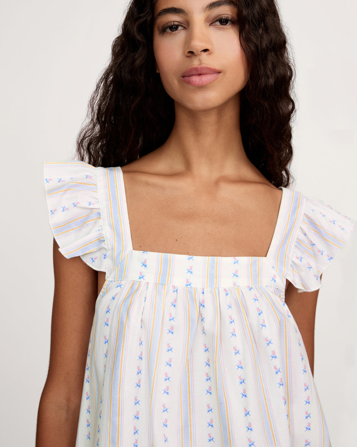 A woman with long curly hair models the Women's Twill Clara Short Set in Cottage Stripe—a loose, sleeveless pajama set with ruffled shoulders and shorts, featuring white fabric with pastel stripes and floral patterns—against a plain background.