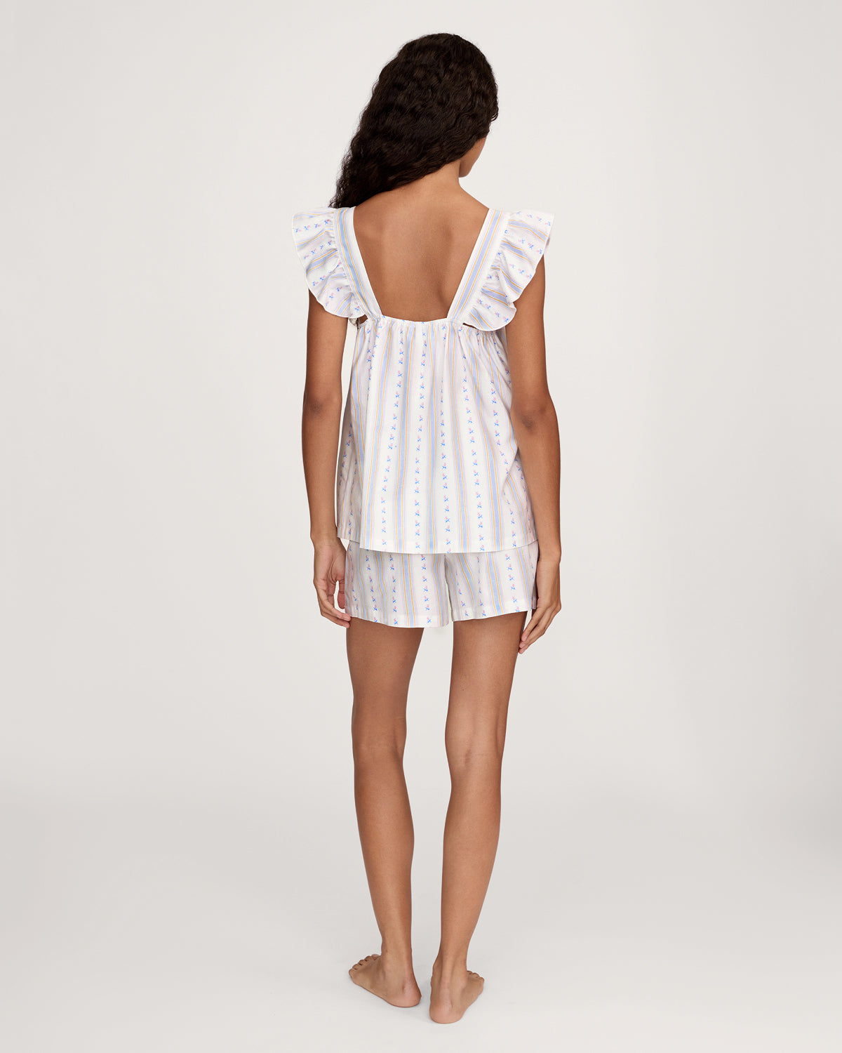 A woman with long curly hair models the Women's Twill Clara Short Set in Cottage Stripe—a loose, sleeveless pajama set with ruffled shoulders and shorts, featuring white fabric with pastel stripes and floral patterns—against a plain background.