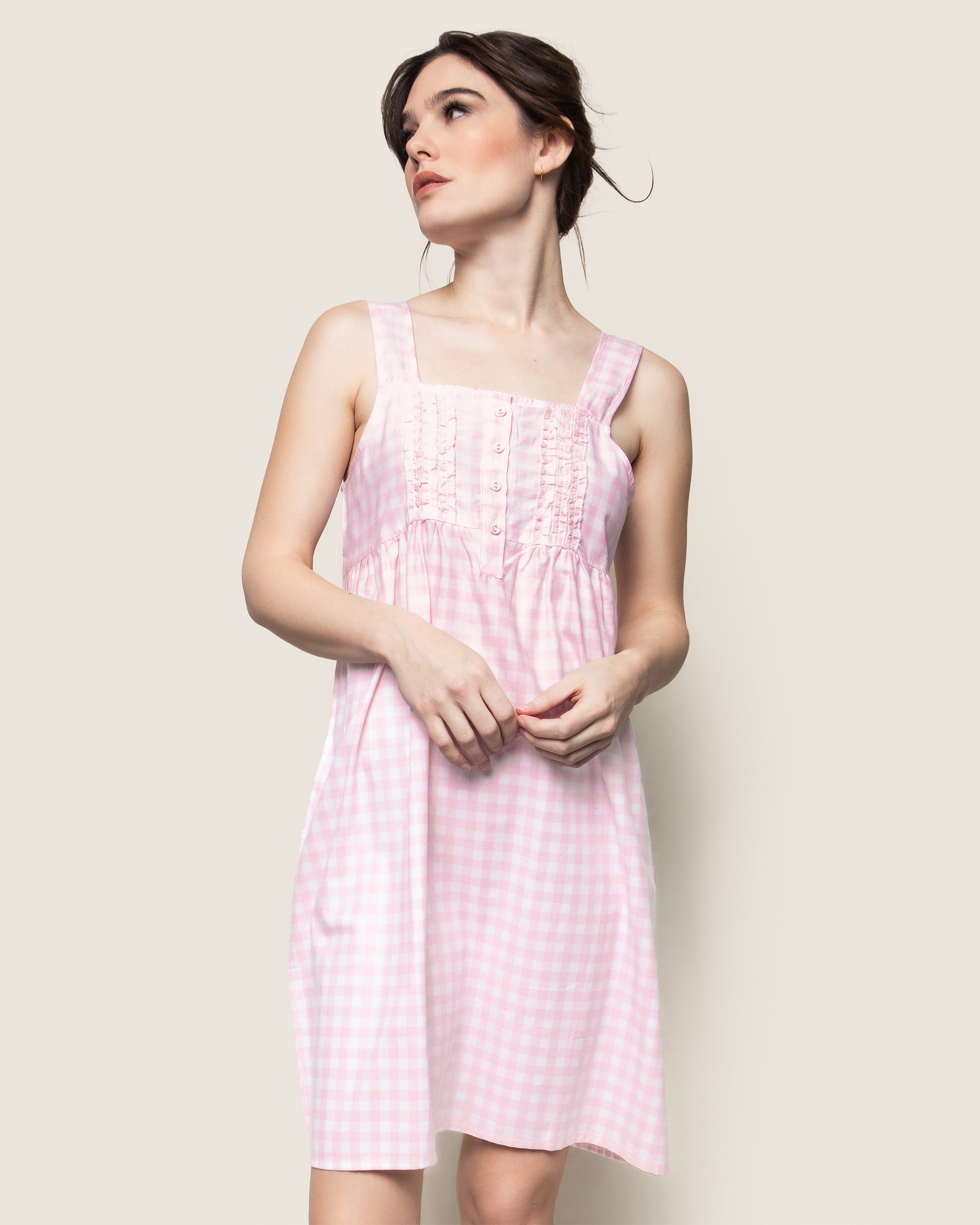 A person in a Petite Plume Womens Twill Charlotte Nightgown in Pink Gingham stands against a plain background, looking to the side with their hands clasped and showcasing a gathered bodice with decorative buttons.
