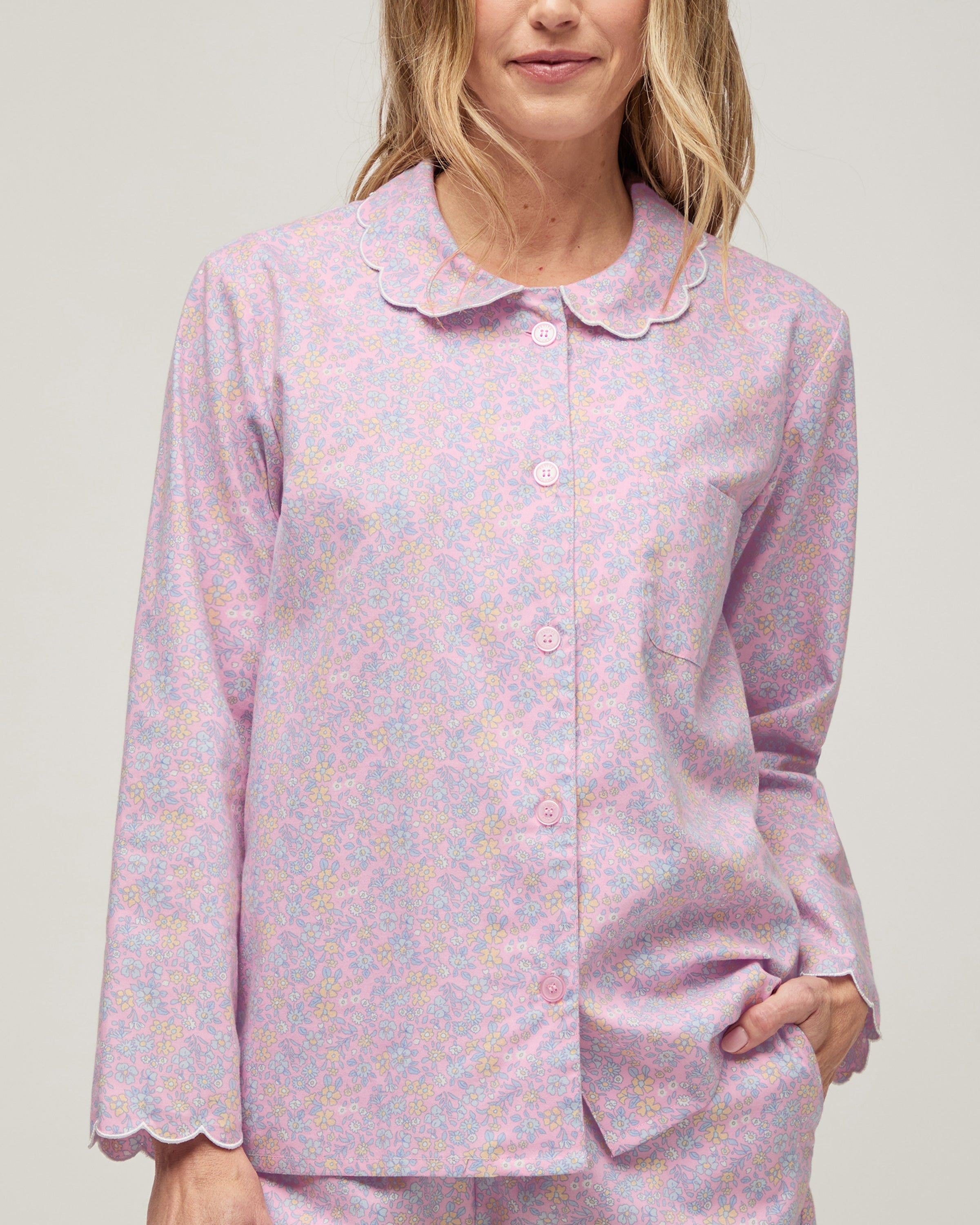 A woman stands barefoot against a plain background, wearing the Women's Twill Colette Pajama Set in Fleur Blush—a matching long-sleeve, button-up top and pants with a light purple and pink floral pattern.