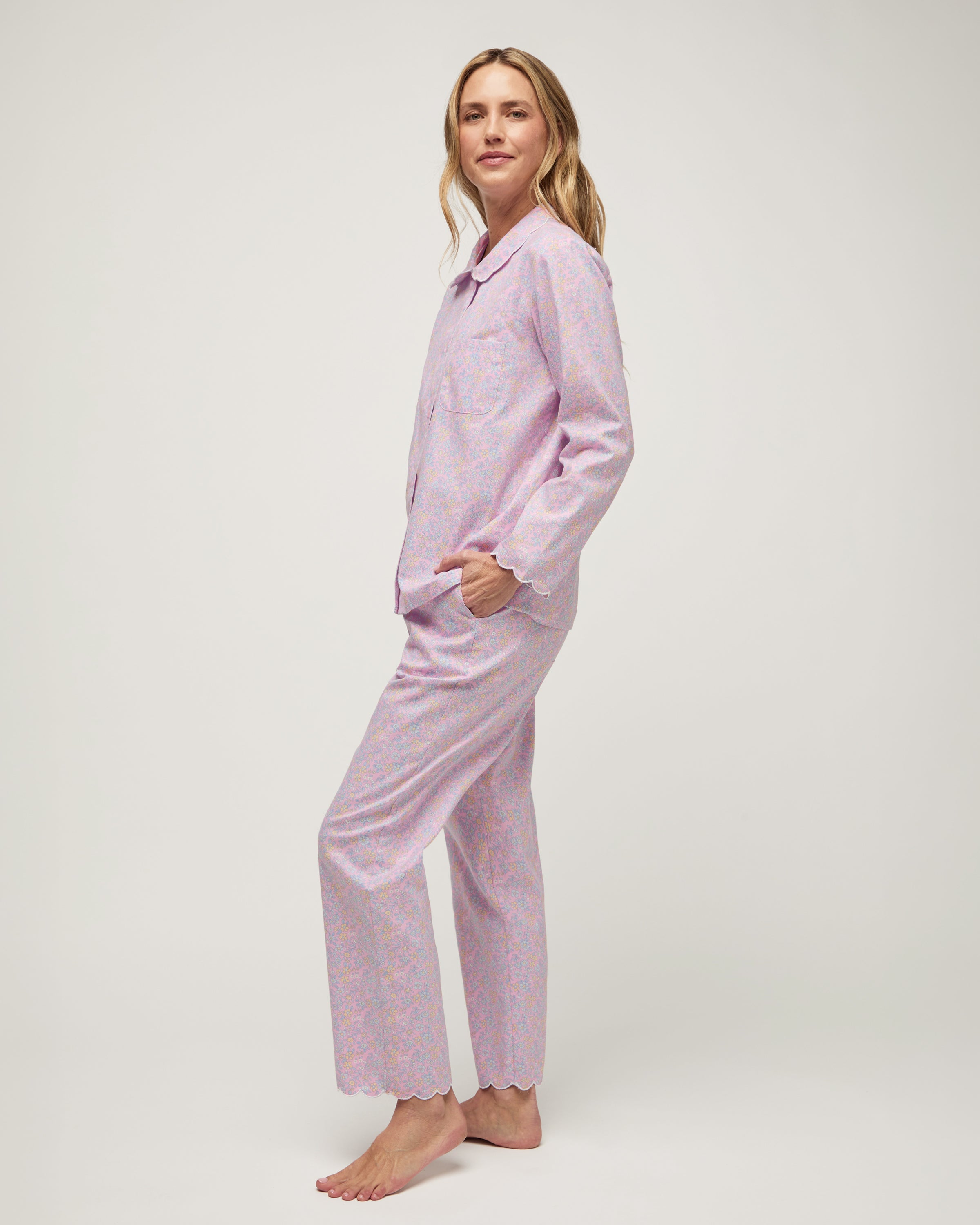 A woman stands barefoot against a plain background, wearing the Women's Twill Colette Pajama Set in Fleur Blush—a matching long-sleeve, button-up top and pants with a light purple and pink floral pattern.