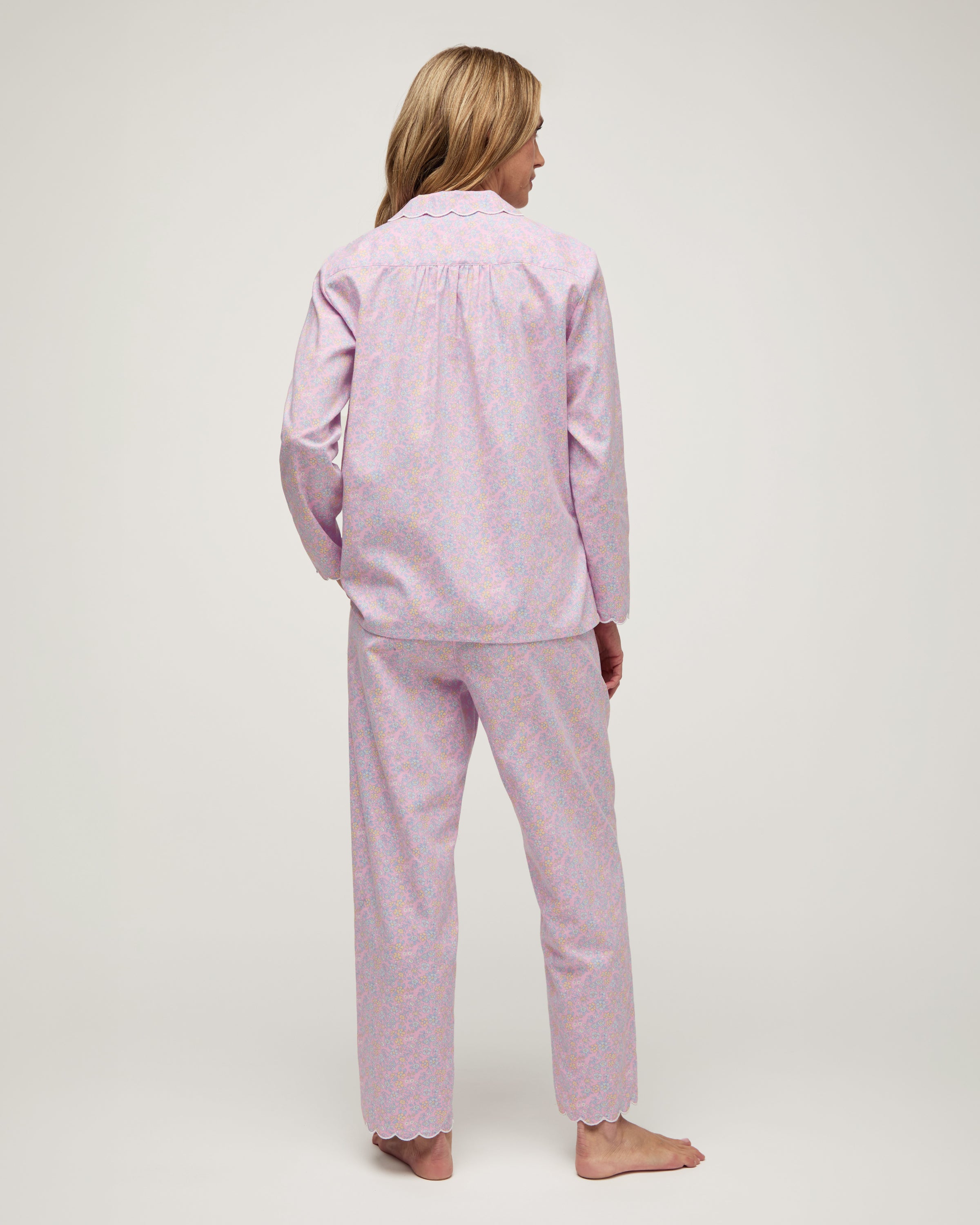 A woman stands barefoot against a plain background, wearing the Women's Twill Colette Pajama Set in Fleur Blush—a matching long-sleeve, button-up top and pants with a light purple and pink floral pattern.