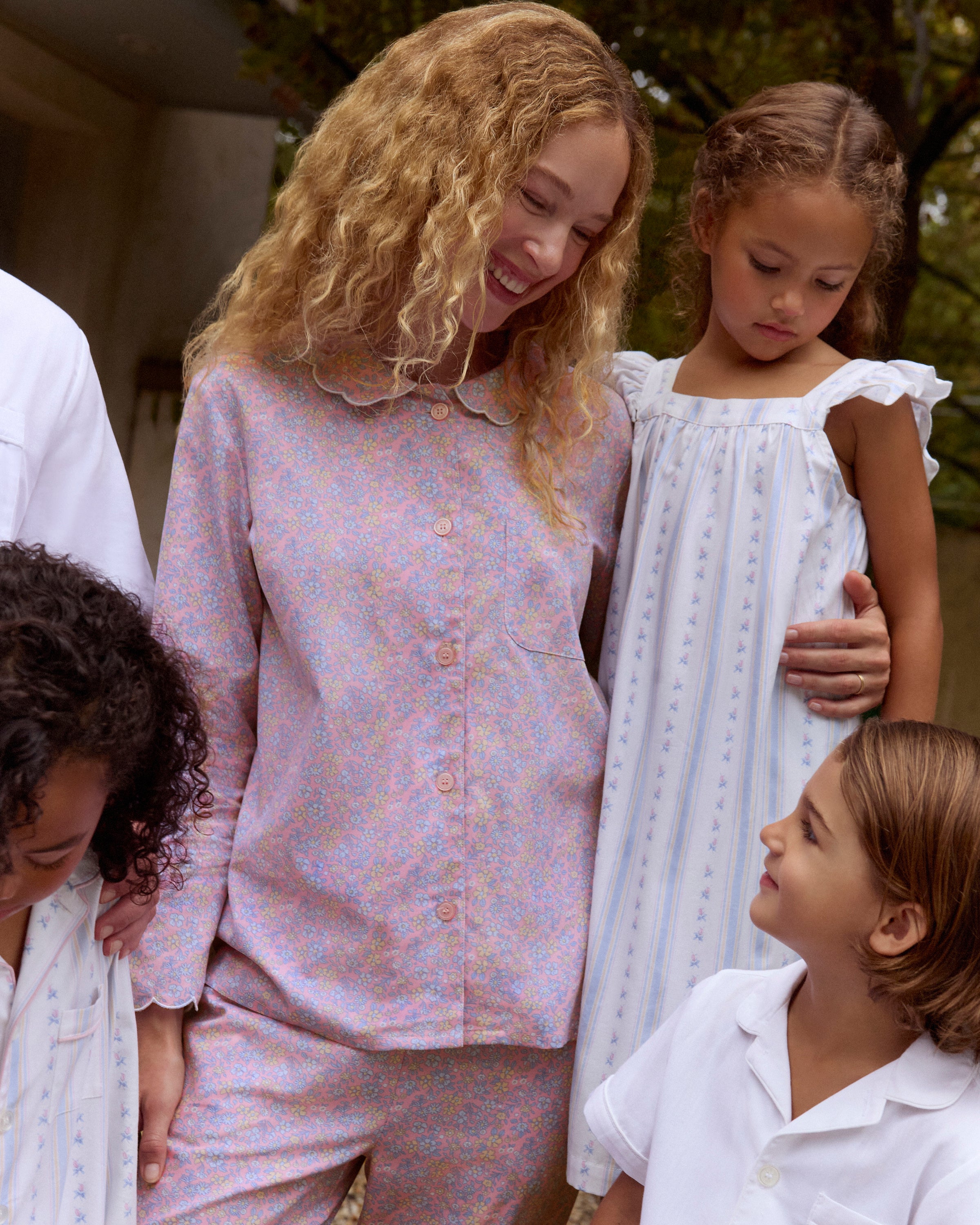 A woman stands barefoot against a plain background, wearing the Women's Twill Colette Pajama Set in Fleur Blush—a matching long-sleeve, button-up top and pants with a light purple and pink floral pattern.