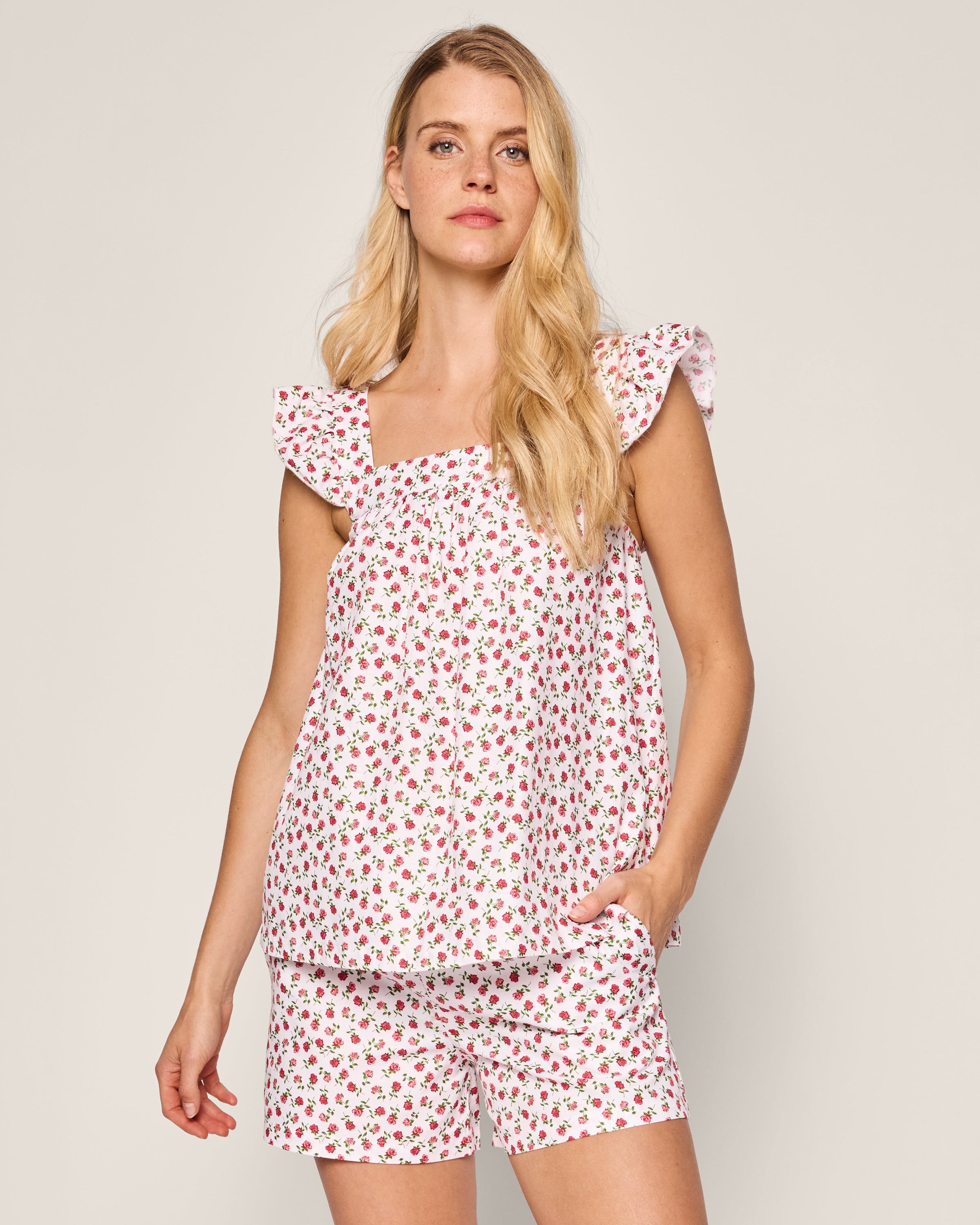 A long-haired blonde is wearing the Womens Twill Clara Short Set in Petite Petals by Petite Plume. The cotton white sleeveless top with ruffled sleeves and a red floral pattern matches the shorts, set elegantly against a plain background, exuding luxury sleepwear vibes.
