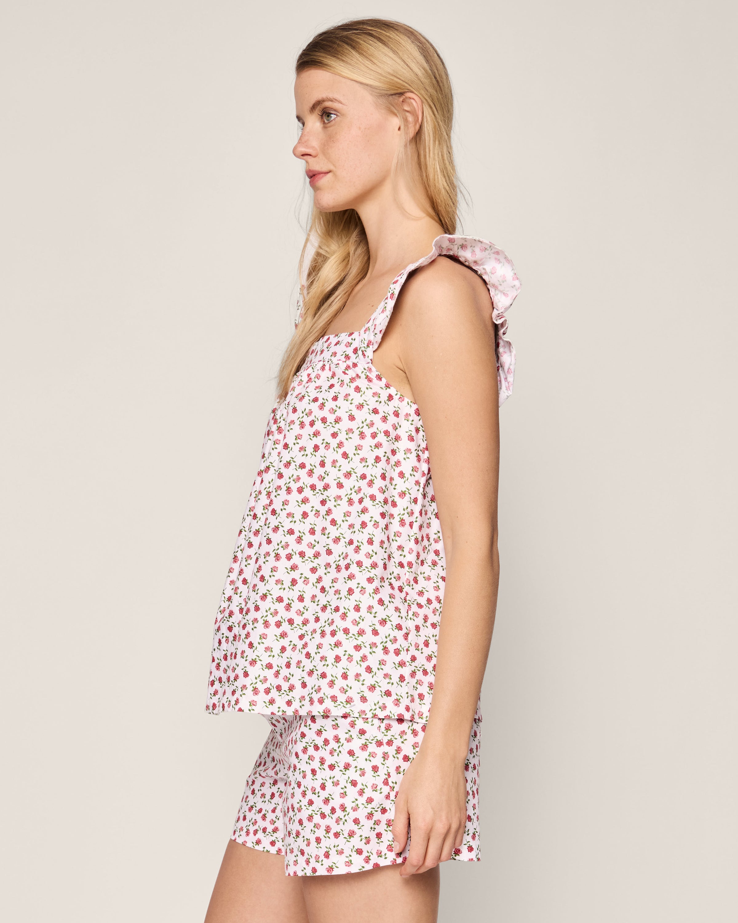 A woman with long blonde hair, in Petite Plumes Womens Twill Clara Short Set in Petite Petals, stands sideways on a light background. The cotton sleeveless top and shorts feature petite red flowers on white with ruffled straps as she gazes away.