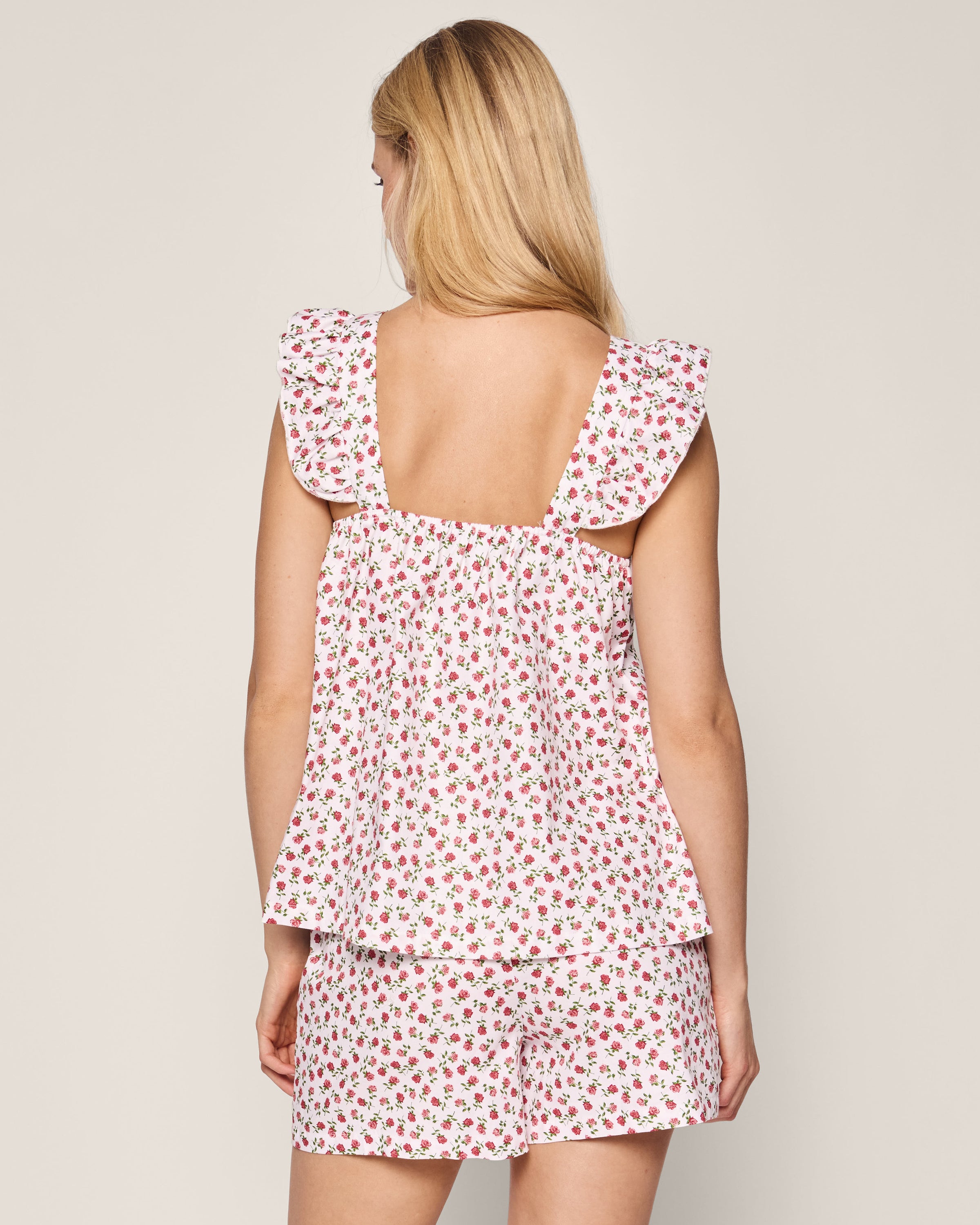 A long-haired blonde woman faces away, wearing the Womens Twill Clara Short Set by Petite Plume. The red and pink floral ensemble includes a sleeveless top with a square neckline and ruffled shoulder straps, crafted from luxurious cotton sleepwear fabric.