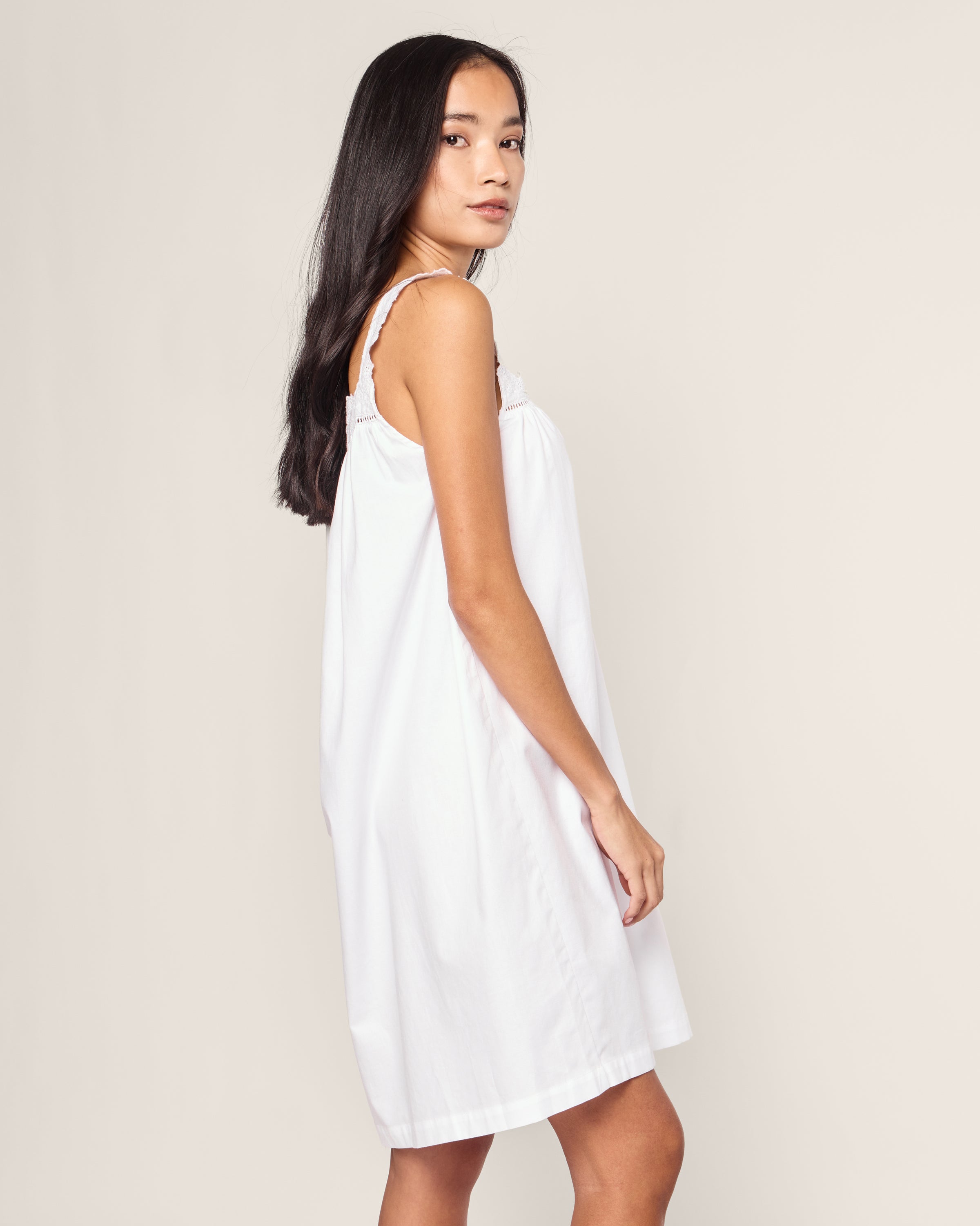 A woman with long dark hair is wearing the elegant Womens Twill Esme Nightgown in White by Petite Plume. She stands sideways, looking over her shoulder against a plain background.