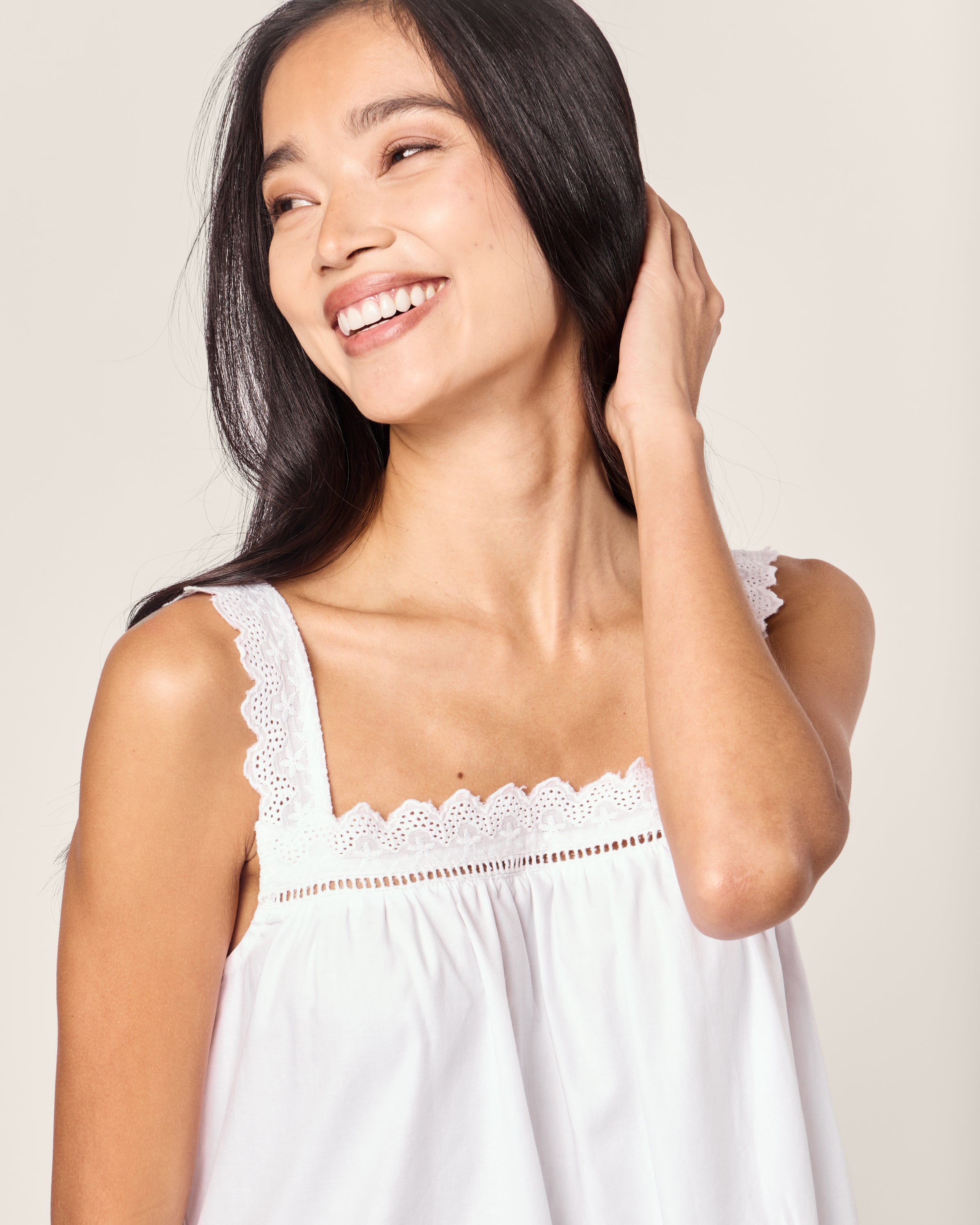 A woman with long black hair smiles while looking left, wearing the luxurious Womens Twill Esme Nightgown in White by Petite Plume. Its sleeveless design features lace detailing on the straps, reminiscent of elegant sleepwear, as she gently touches her hair against a light neutral background.