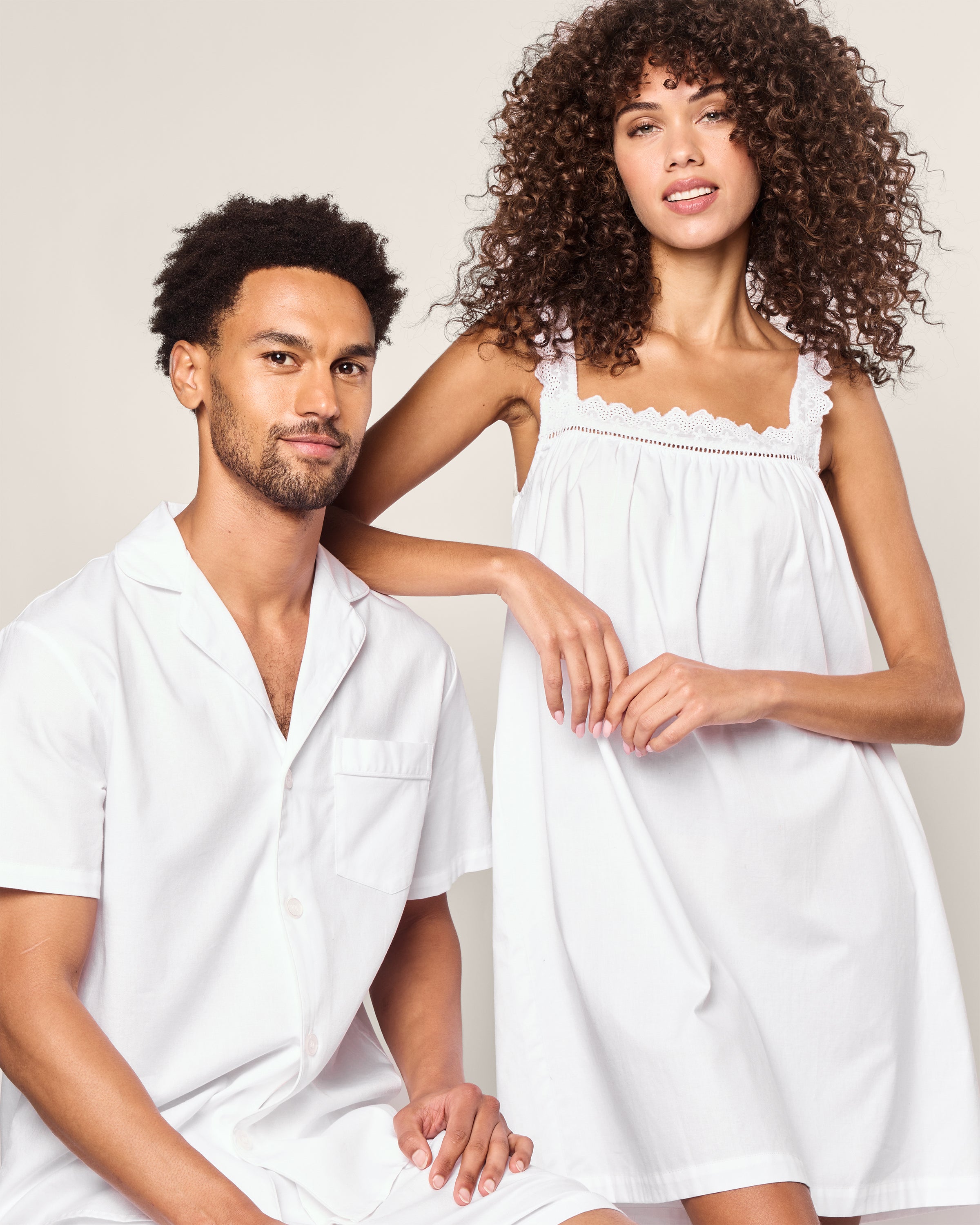 A man in a white button-up pajama shirt and shorts sits comfortably, while a woman in Petite Plumes Womens Twill Esme Nightgown in White stands gracefully beside him with her arm on his shoulder. Both wear luxurious cotton sleepwear, and the nightgown features elegant lace trim.
