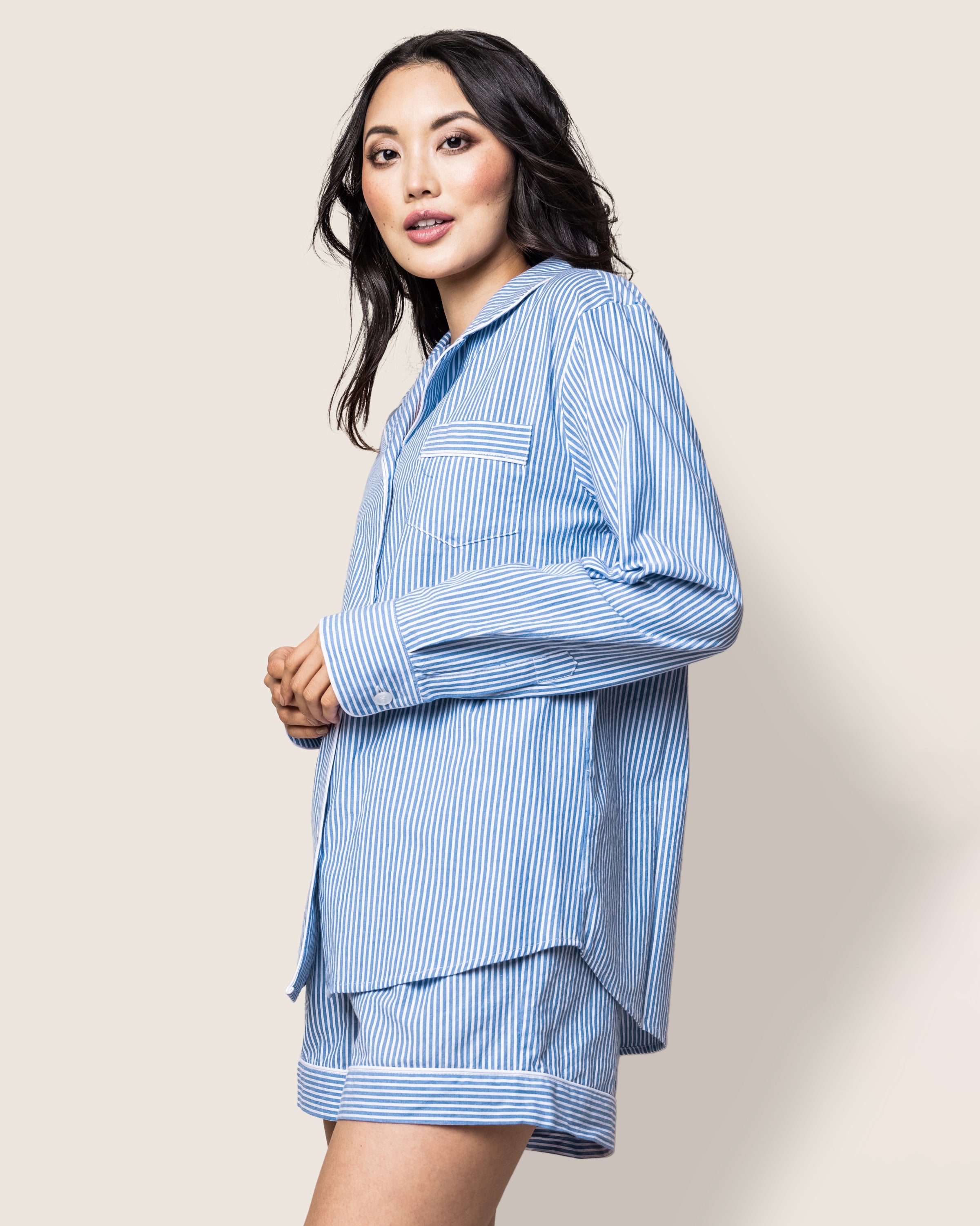 A person with long hair wears the luxurious Womens Twill Long Sleeve Short Set in French Blue Seersucker by Petite Plume, featuring a button-up shirt and shorts. They gaze at the camera with a neutral expression against a plain, light-colored background.