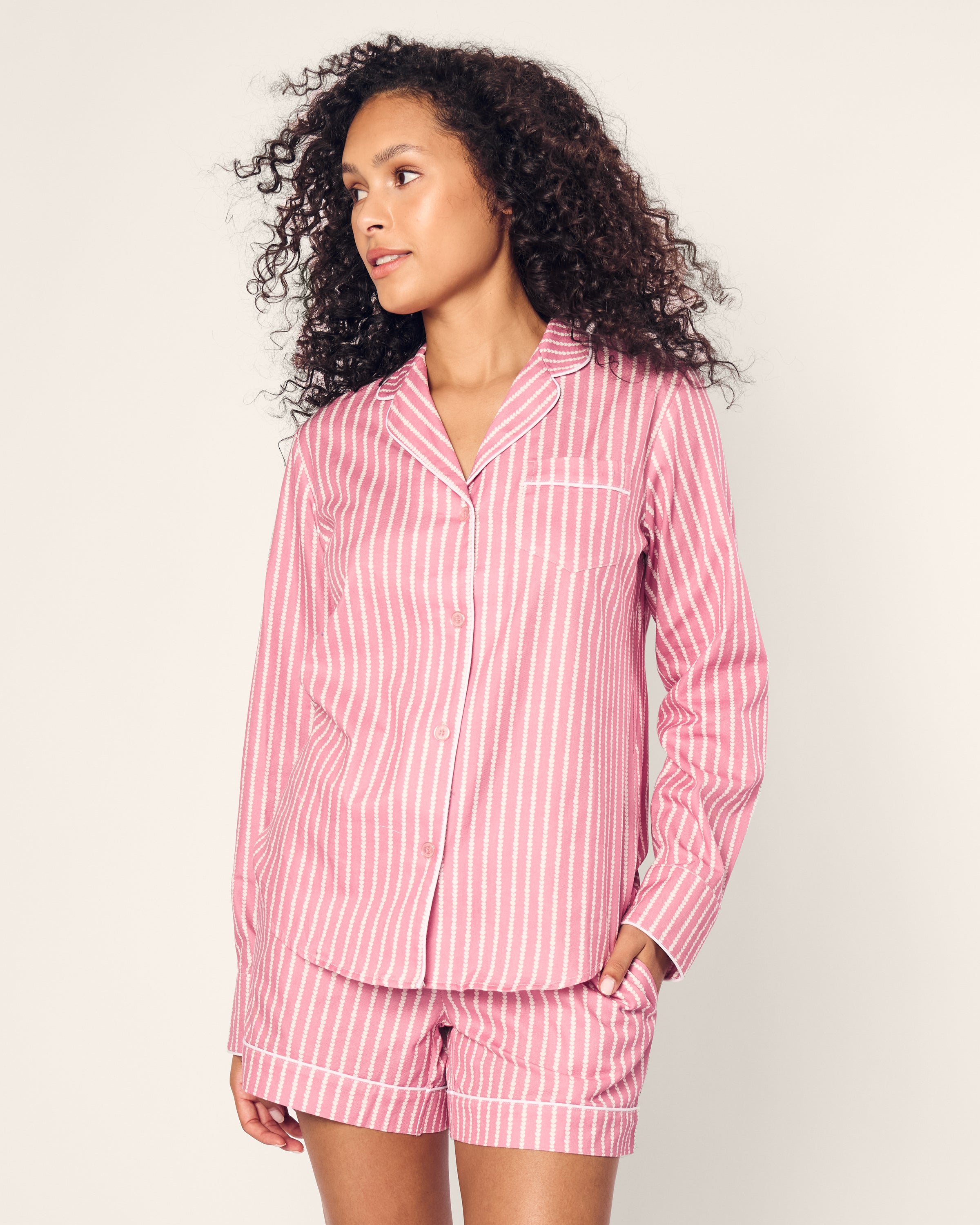 A curly-haired person wears the Womens Twill Long Sleeve Short Set in Love Lines by Petite Plume, featuring a luxurious pink and white vertical striped cotton pajama set with a long-sleeved top and shorts, as they gaze to the side against a plain background.