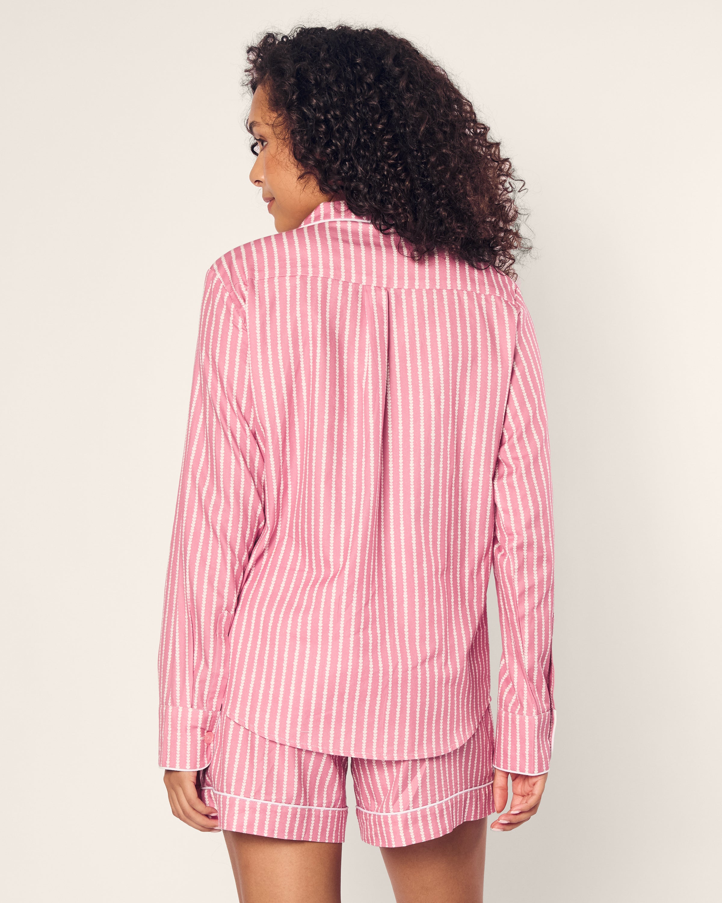 A person with curly hair wears the luxurious Petite Plume Womens Twill Long Sleeve Short Set in Love Lines, featuring pink and white vertical stripes. Made from premium cotton, this sleepwear adds elegance as they stand with their back to the camera against a plain background.