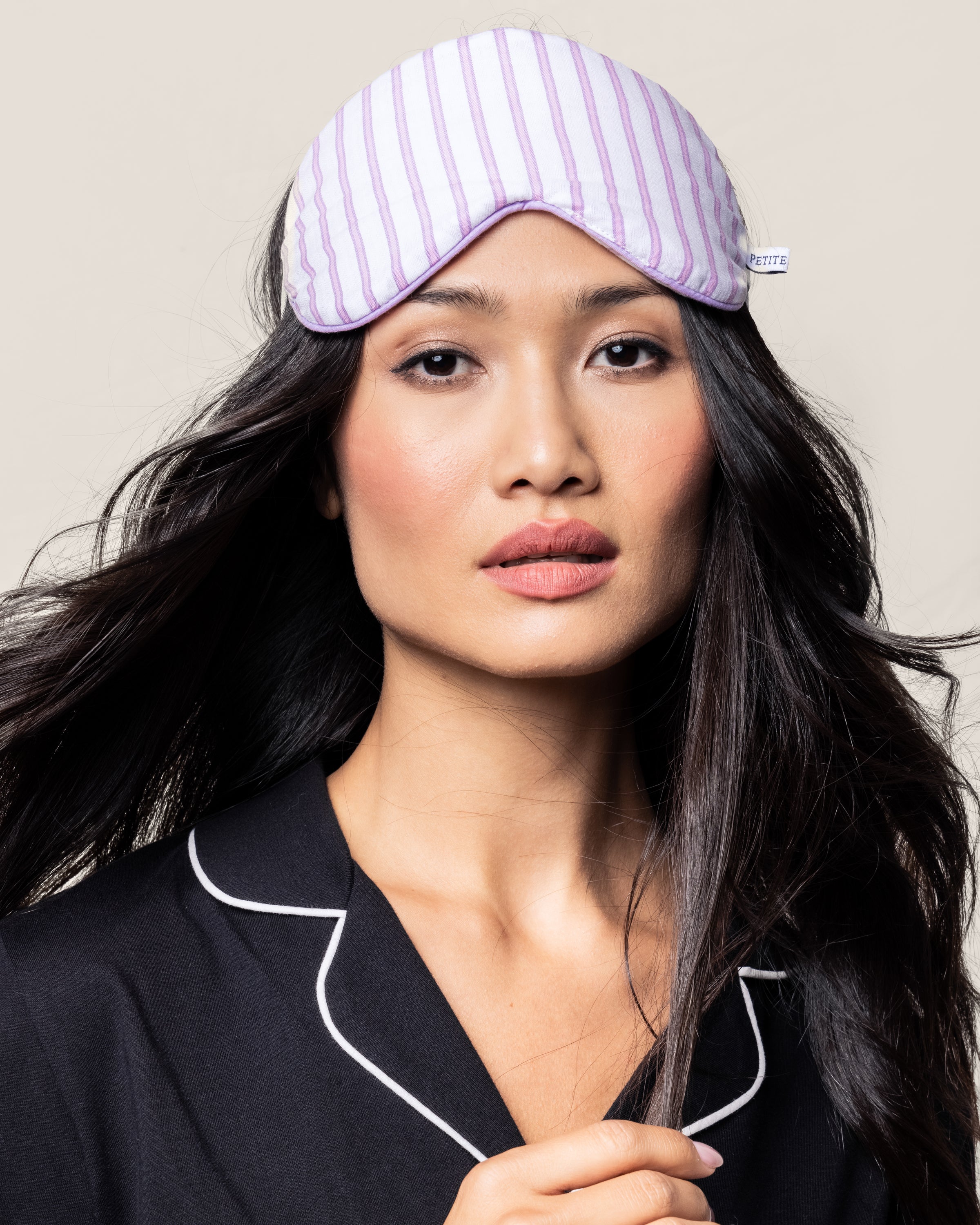 A person with long dark hair wears a monogrammed Petite Plume Adults Twill Sleep Mask in Lavender French Ticking atop their head. Donning a black pajama top with white piping, they stand against a plain beige background.