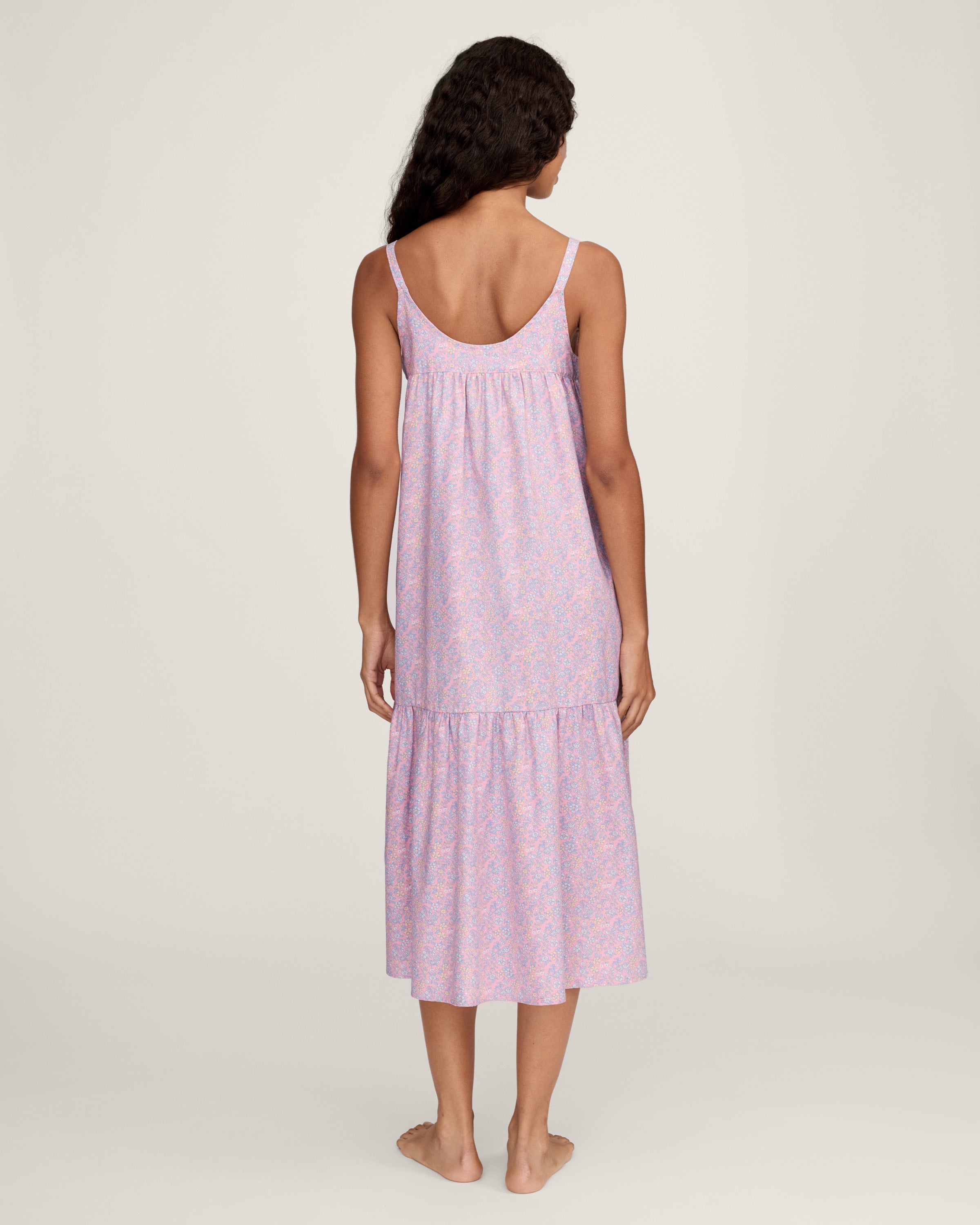 A woman with long curly hair models the Women's Twill Chloé Nightgown in Fleur Blush, a sleeveless, loose-fitting light purple floral dress. She is barefoot and stands relaxed against a plain, light background.