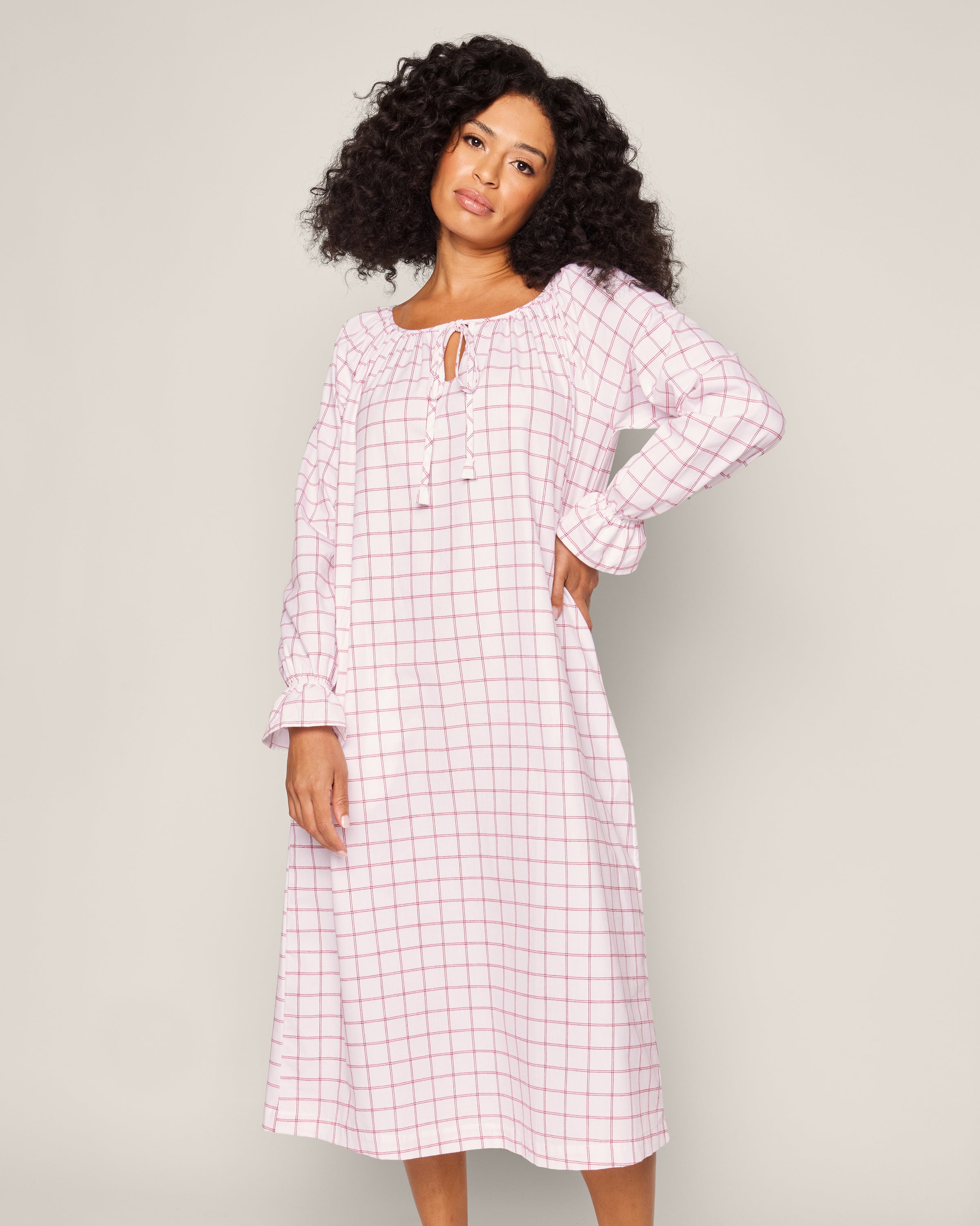 A person with curly hair wears the Womens Twill Delphine Nightgown in Garnet Tattersall by Petite Plume. Featuring a round neckline with a keyhole opening, the white and pink checkered gown exudes confidence against a light gray background.