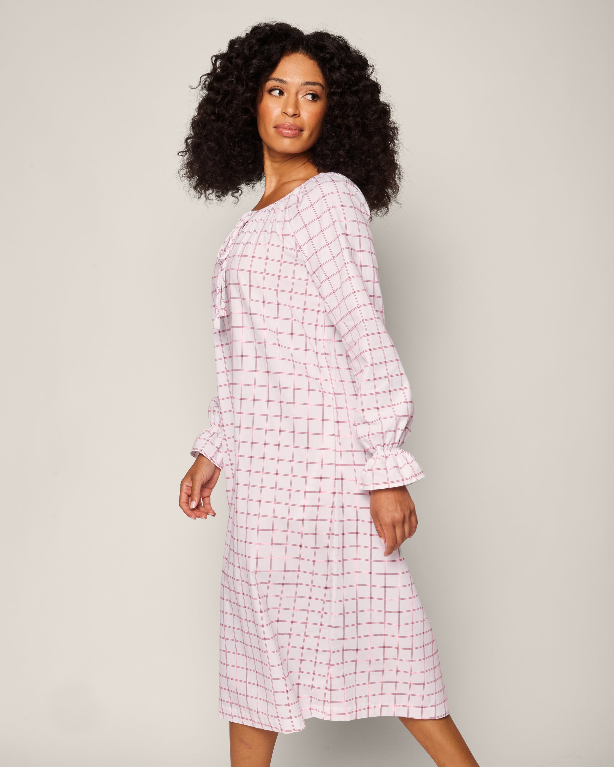 A curly-haired woman wears a Petite Plume Womens Twill Delphine Nightgown in Garnet Tattersall, looking sideways. The cotton twill gown has ruffled sleeve cuffs and a tie detail at the neckline against a plain, neutral background.