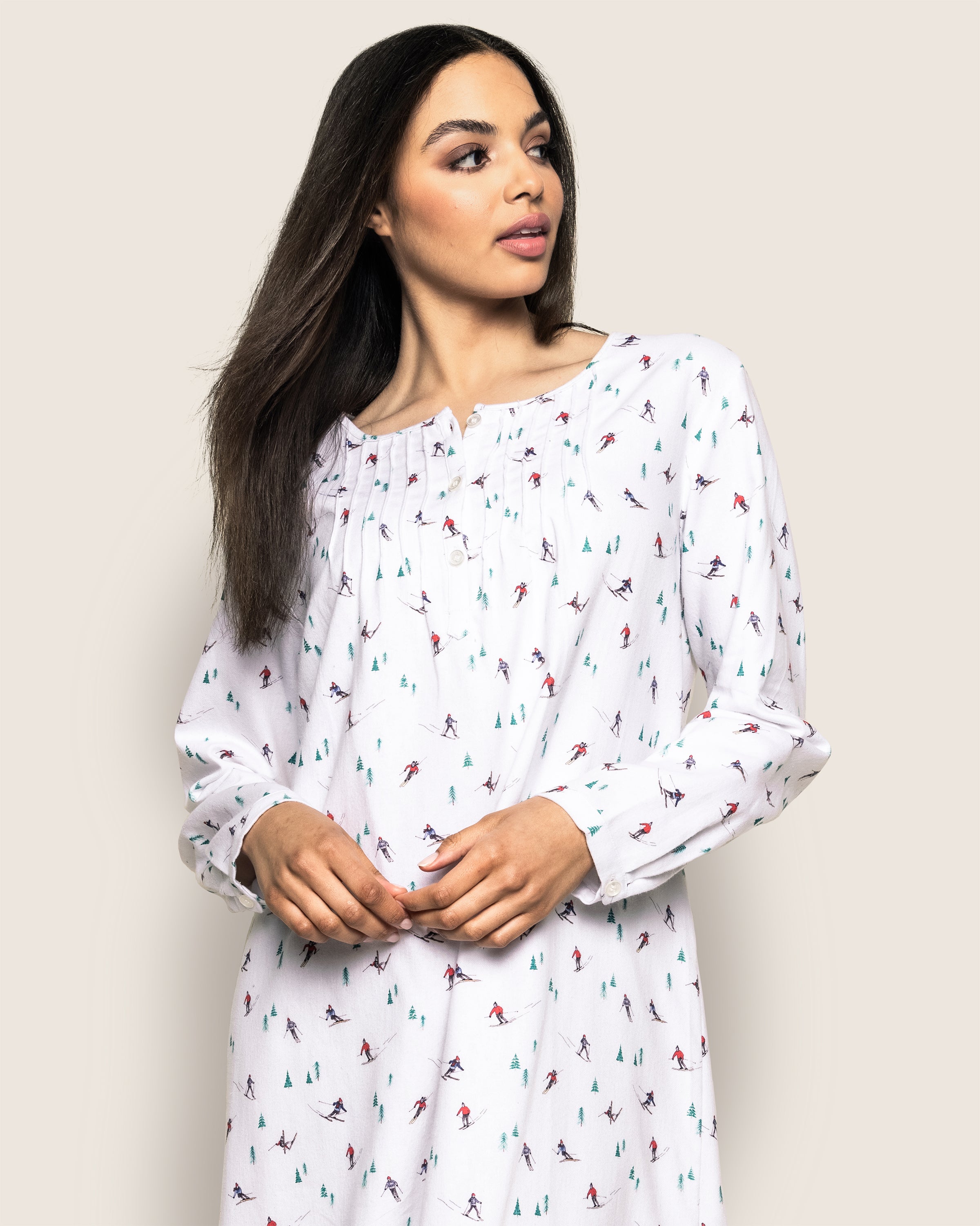 A woman in Petite Plumes Womens Flannel Beatrice Nightgown in Après Ski, adorned with a vibrant skier pattern, looks to the side. Her long dark hair cascades elegantly, while the gowns gathered neckline and long sleeves over a plain light background exude luxurious sleepwear vibes.