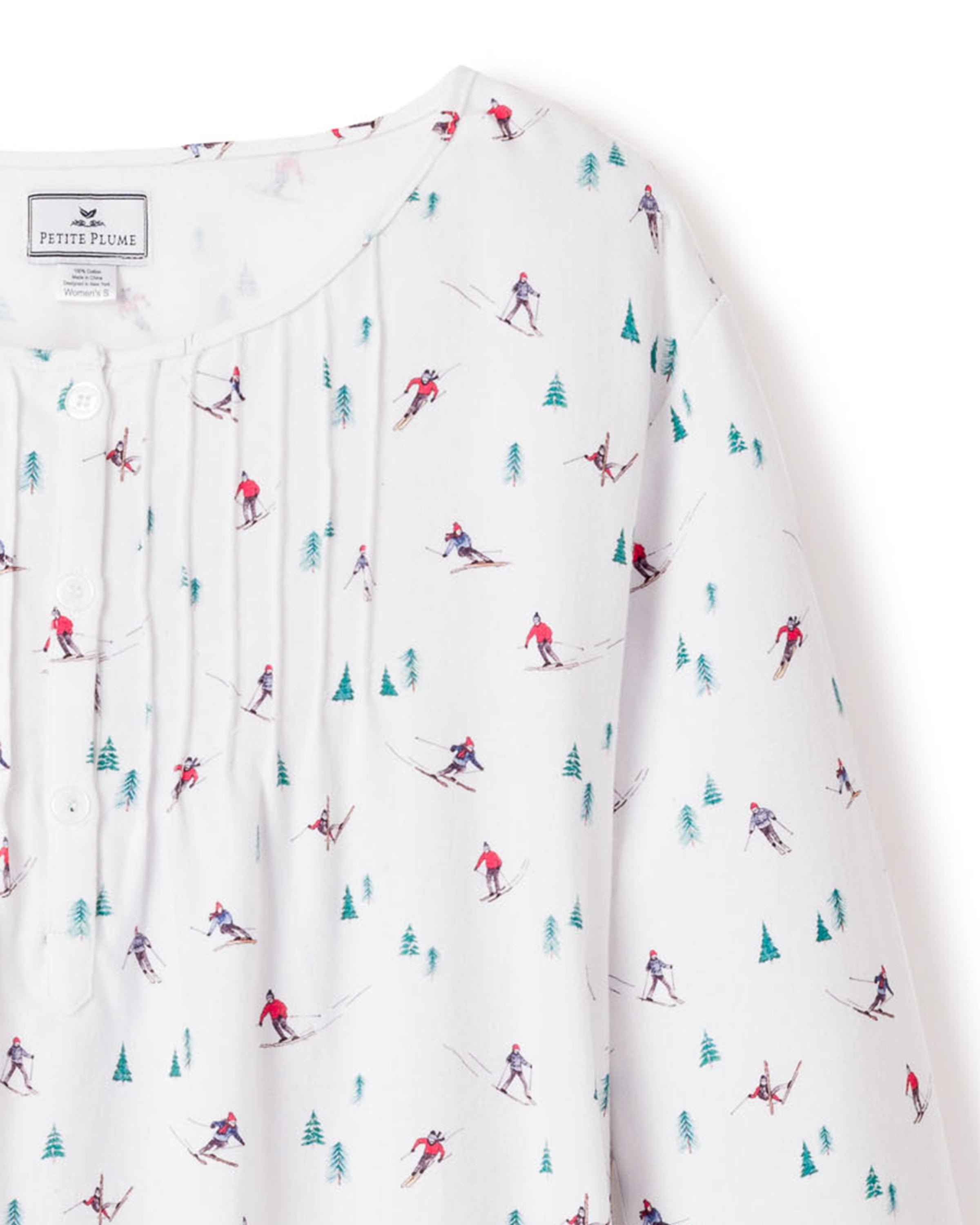 The Womens Flannel Beatrice Nightgown in Après Ski by Petite Plume showcases a white cotton fabric adorned with skiers in red and blue gear among green trees. It features a neckline label and front buttons, highlighting its luxury sleepwear status.