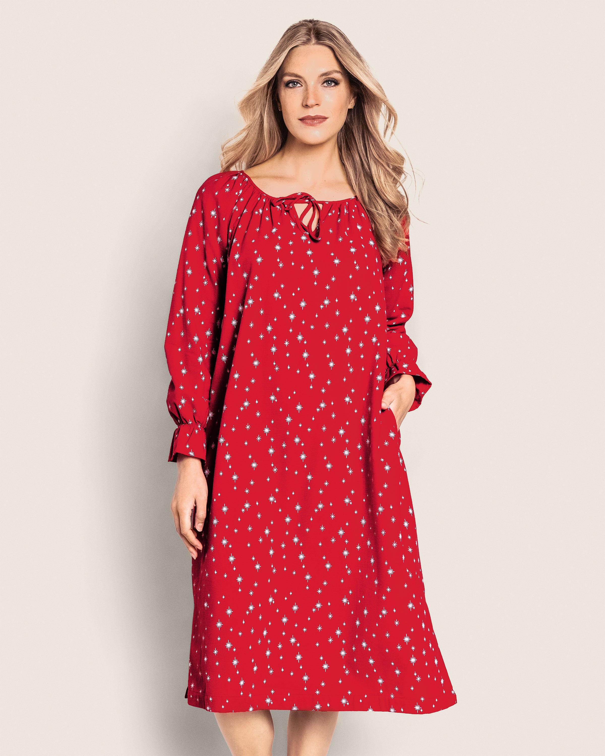 A woman stands against a plain background wearing the Petite Plume Womens Flannel Delphine Nightgown in Starry Night, a loose red dress with white star patterns. The elegant gown features long sleeves, pockets, and complements her long, wavy hair and neutral expression.
