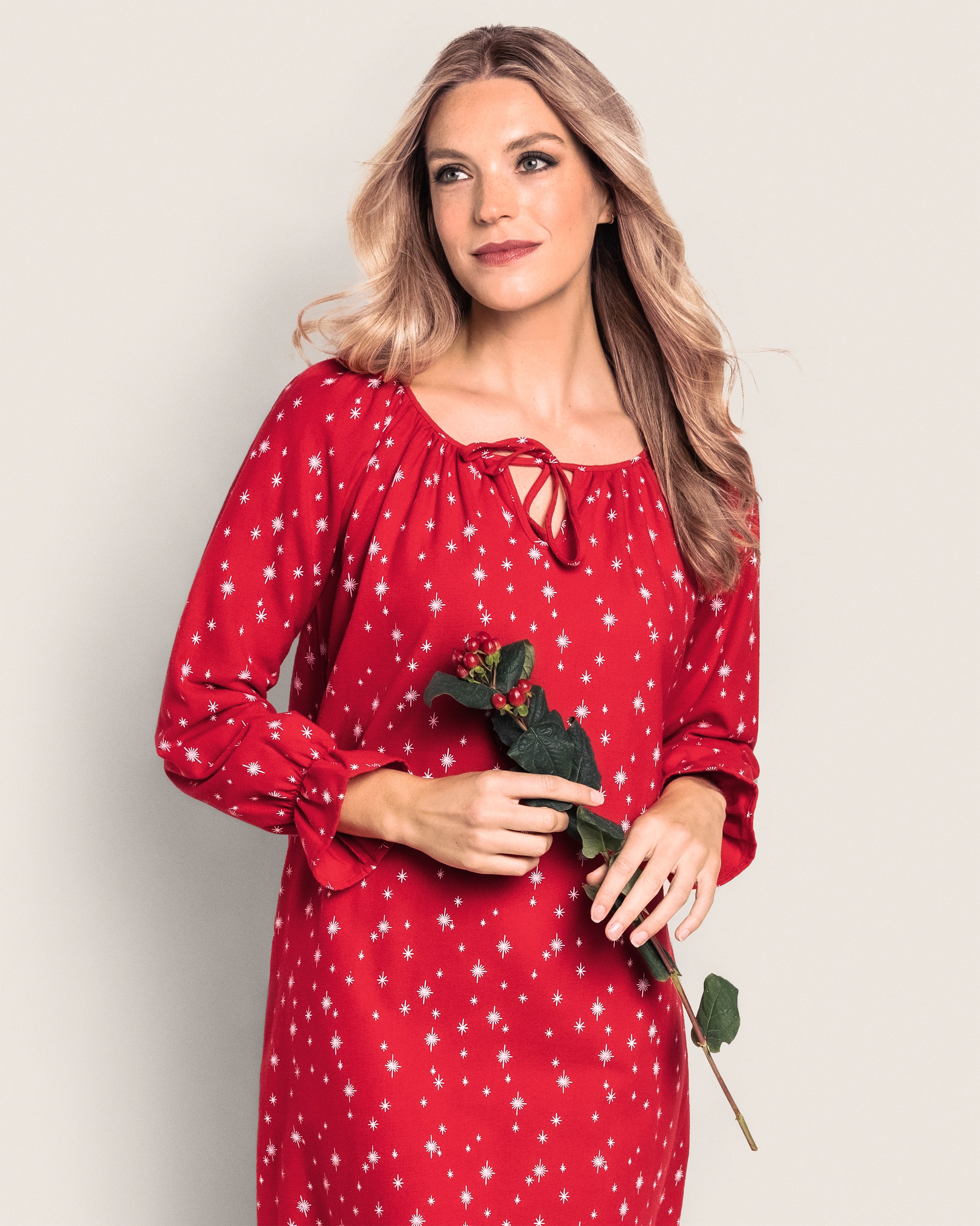 A woman with long blonde hair wears a Womens Flannel Delphine Nightgown in Starry Night by Petite Plume, made from lightweight 100% cotton, as she holds a rose. She gazes slightly to the side, with a gentle smile against a plain light background.