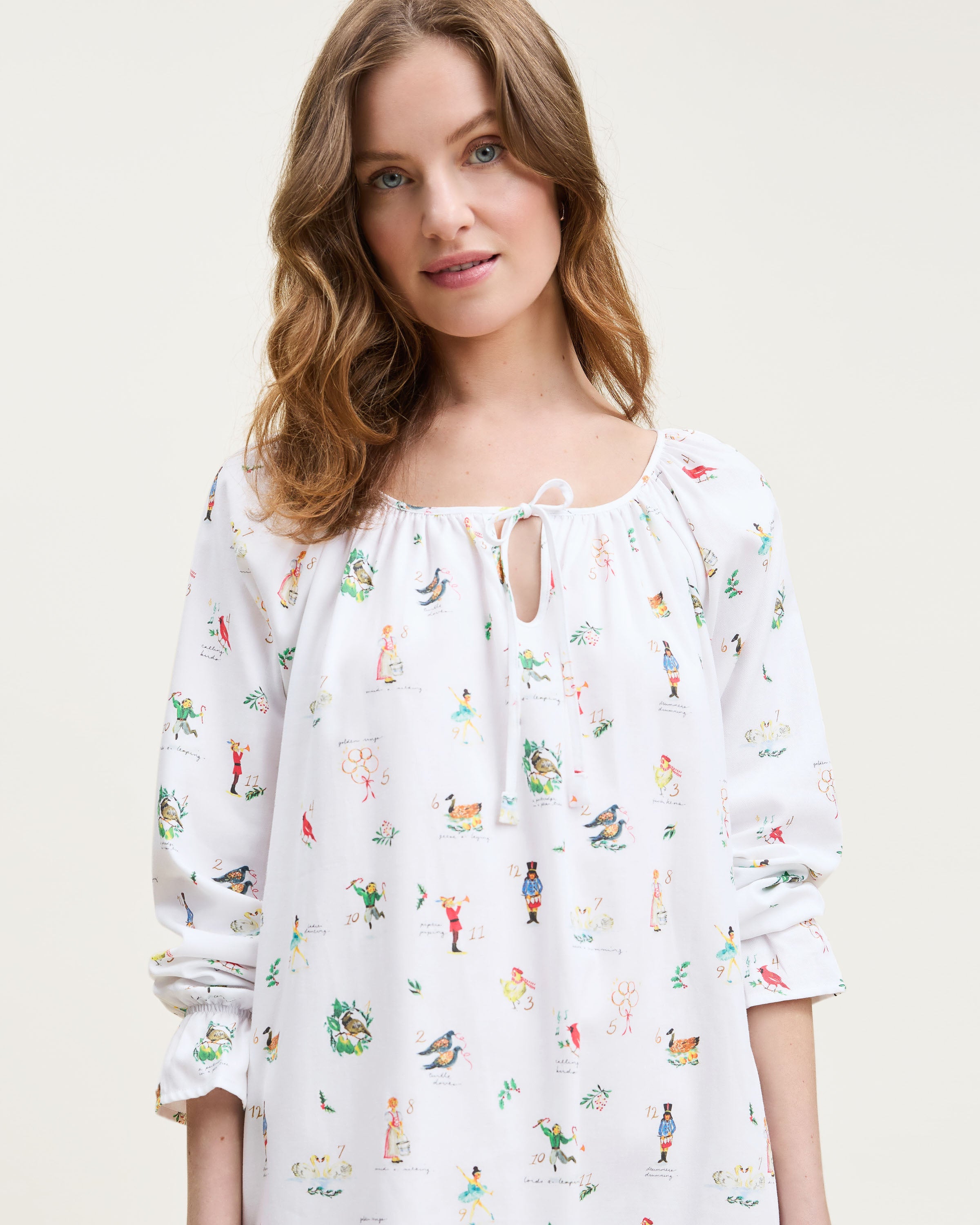The Women's Twill Delphine Nightgown in 12 Days of Christmas is a white, long-sleeve nightgown with a gathered neckline, small bow tie, and a festive print inspired by the 12 Days of Christmas.