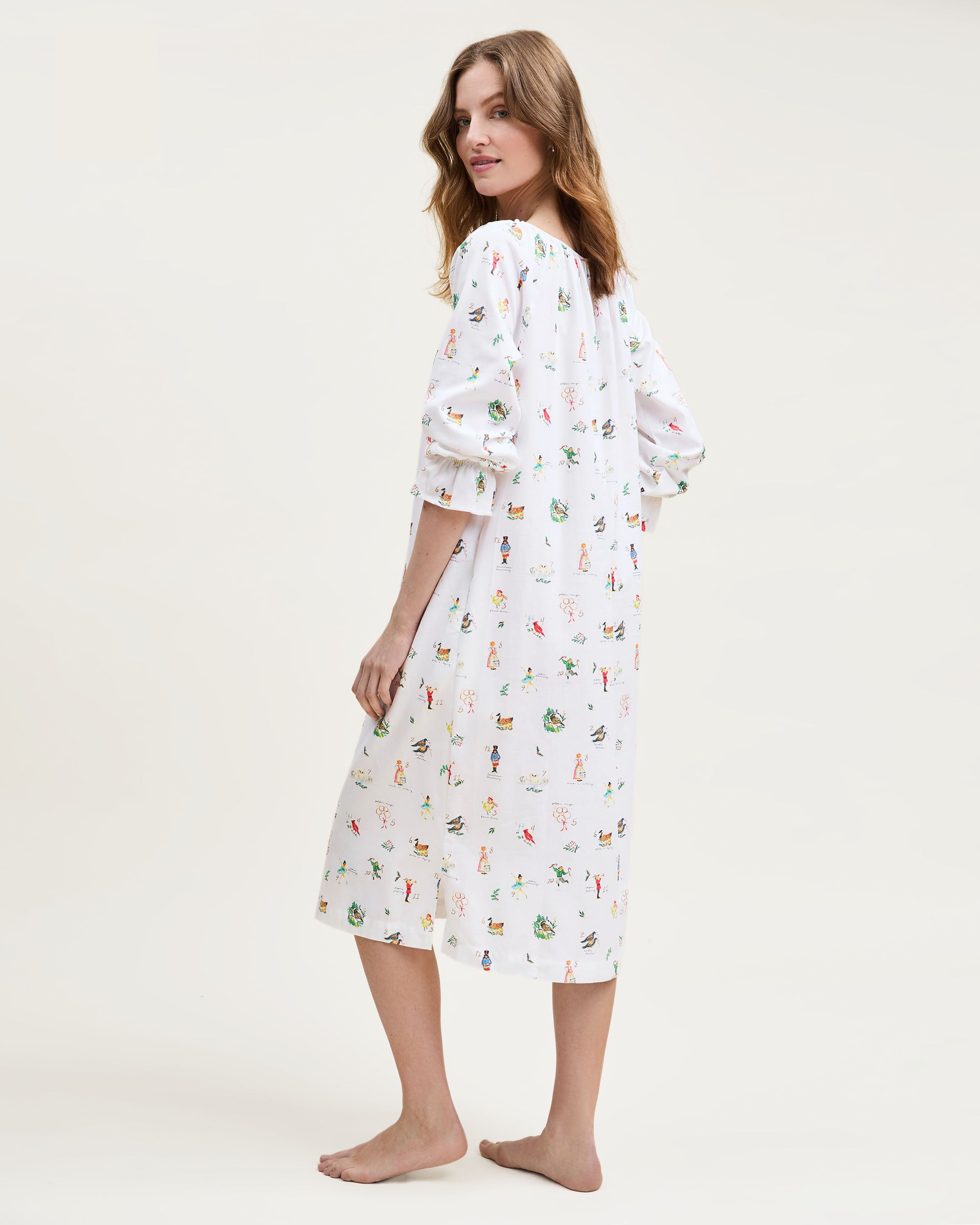 The Women's Twill Delphine Nightgown in 12 Days of Christmas is a white, long-sleeve nightgown with a gathered neckline, small bow tie, and a festive print inspired by the 12 Days of Christmas.