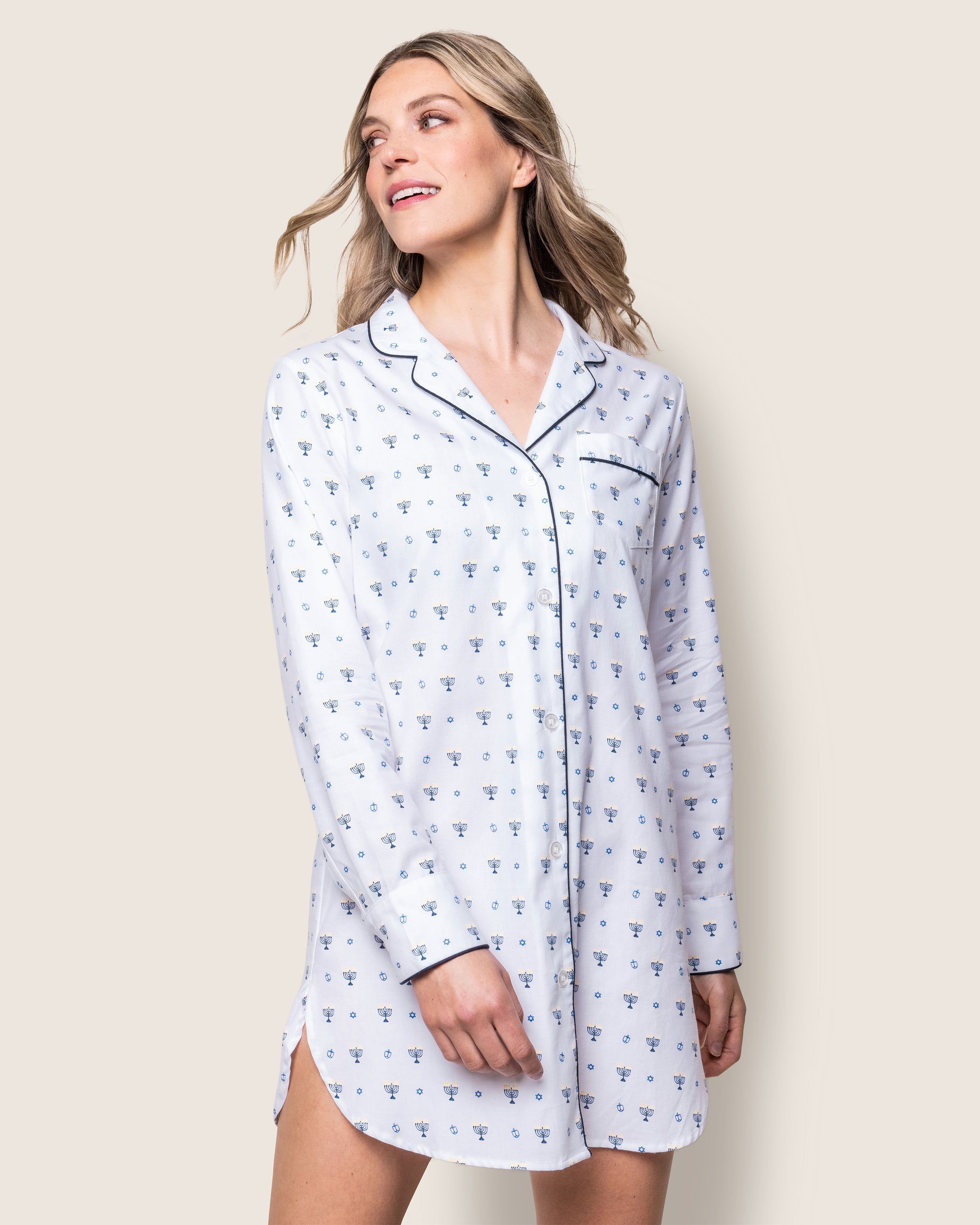 Against a light backdrop, a person with long, wavy hair in a Womens Twill Nightshirt in Happy Hanukkah by Petite Plume smiles gently. The white cotton nightshirt with blue patterns features a collar and dark piping along the edges, inspired by Hanukkah pajamas.