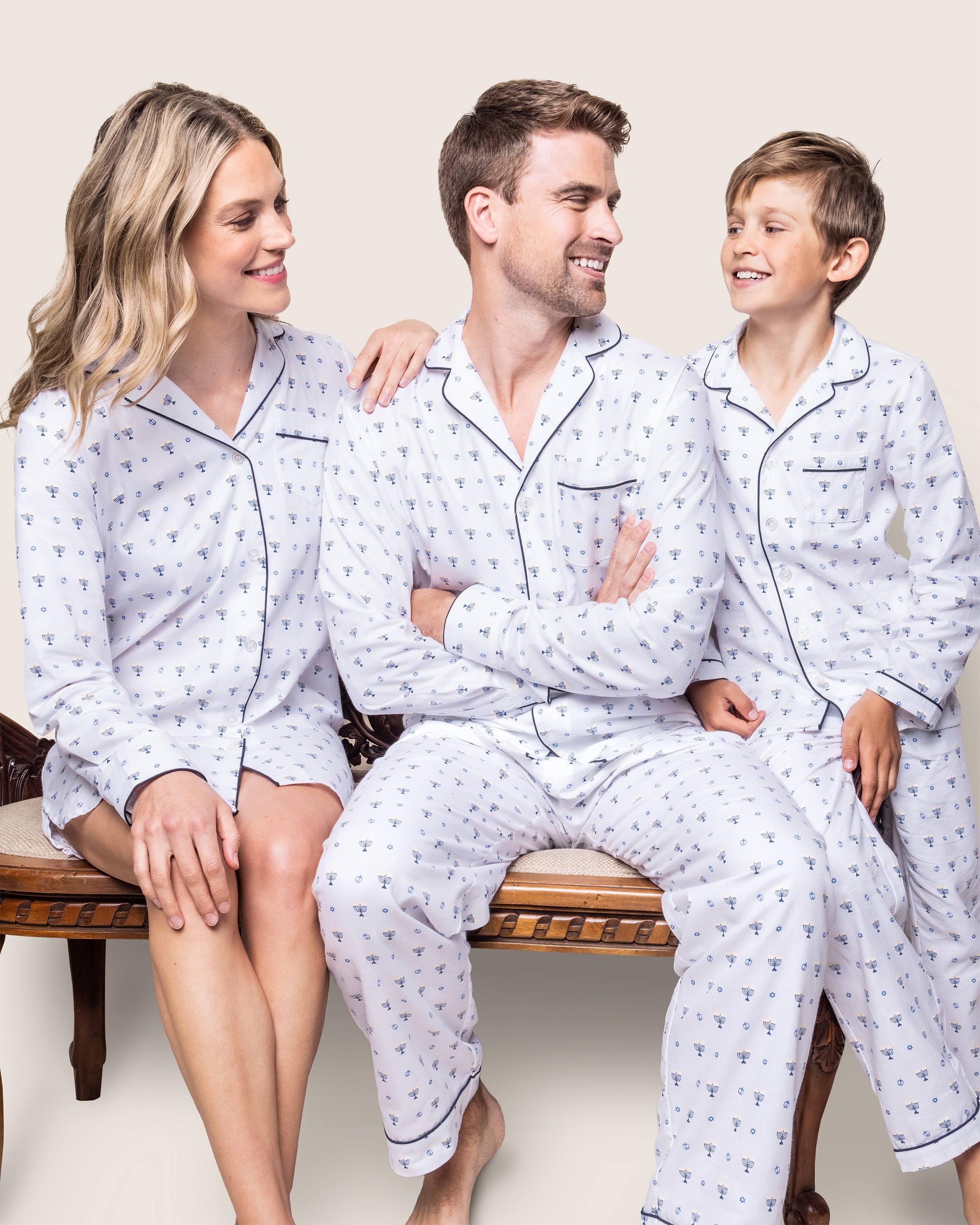 A family of three sits on a bench in matching Petite Plume nightwear. The adults, wearing Womens Twill Nightshirt in Happy Hanukkah design, are on either side of the child. They exchange warm smiles against a plain backdrop, celebrating Hanukkah together.
