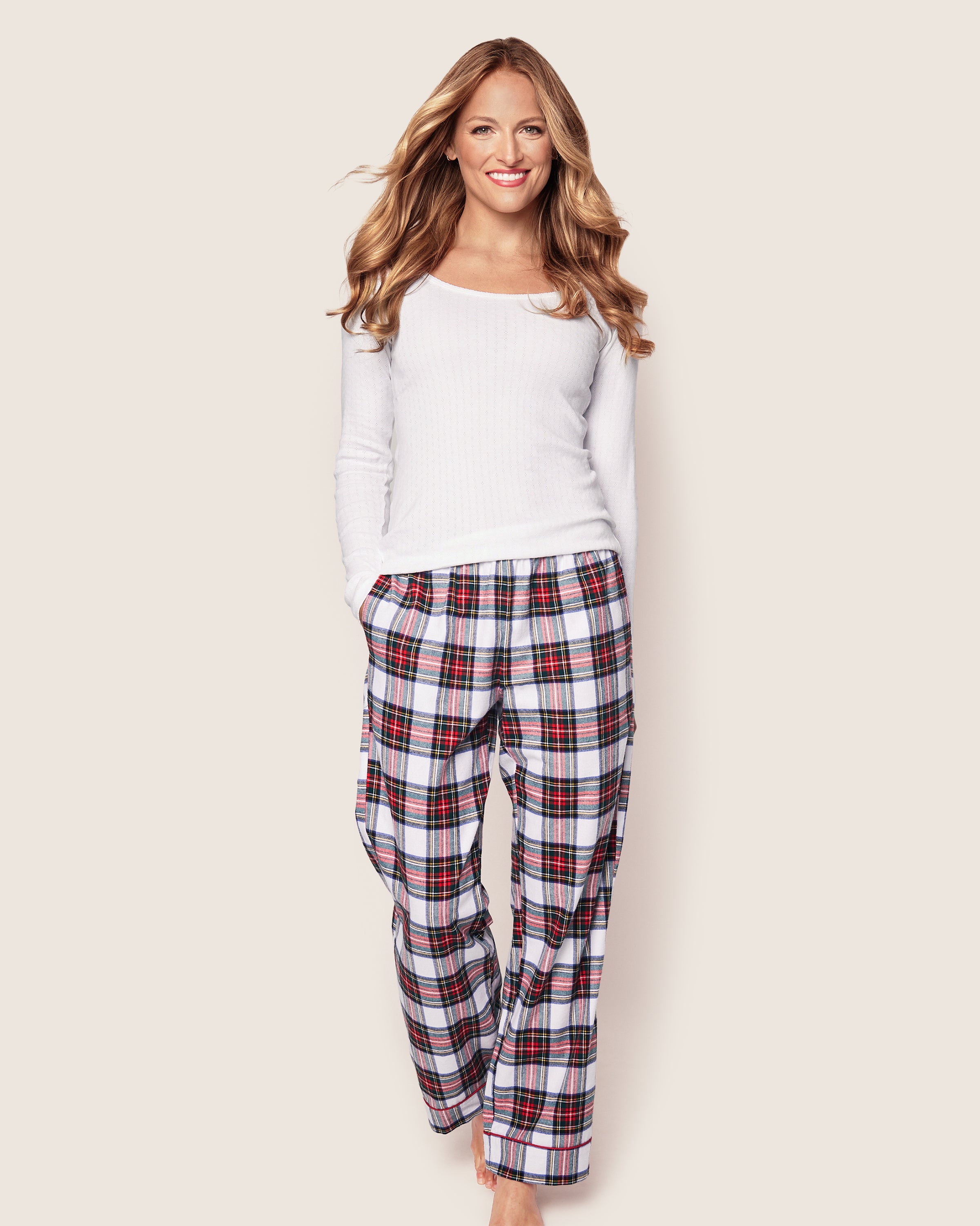 A woman with long, wavy hair wears a luxurious white top and Petite Plumes Womens Brushed Cotton Pants in Balmoral Tartan. She stands barefoot against a light background, smiling with her hands in her pockets.
