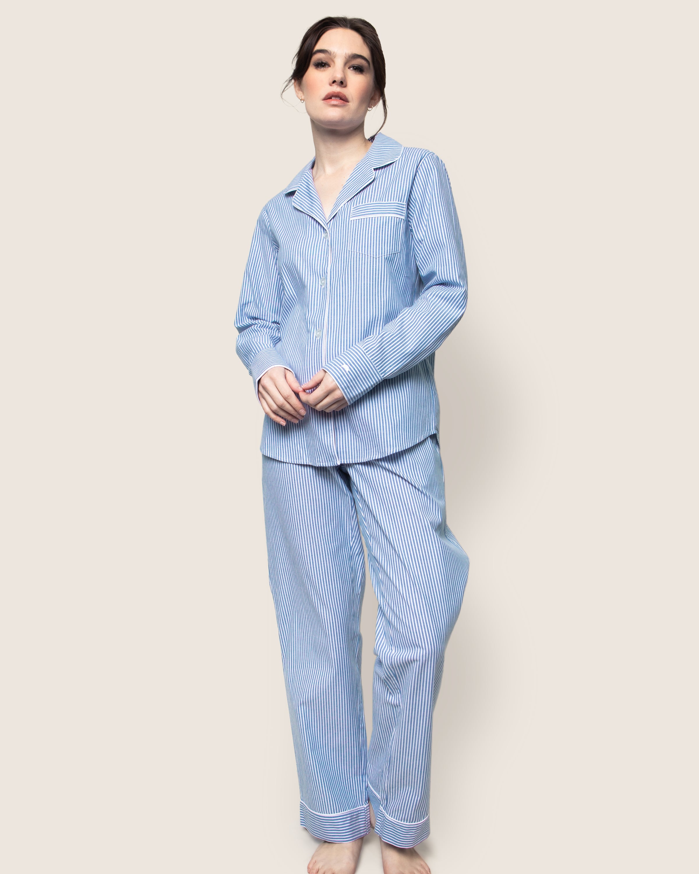 A person in a Petite Plume Womens Twill Pajama Set, featuring French Blue Seersucker 100% cotton fabric, stands barefoot against a plain background. The classic long-sleeve button-up top is paired with matching pants, and their hair is tied back.