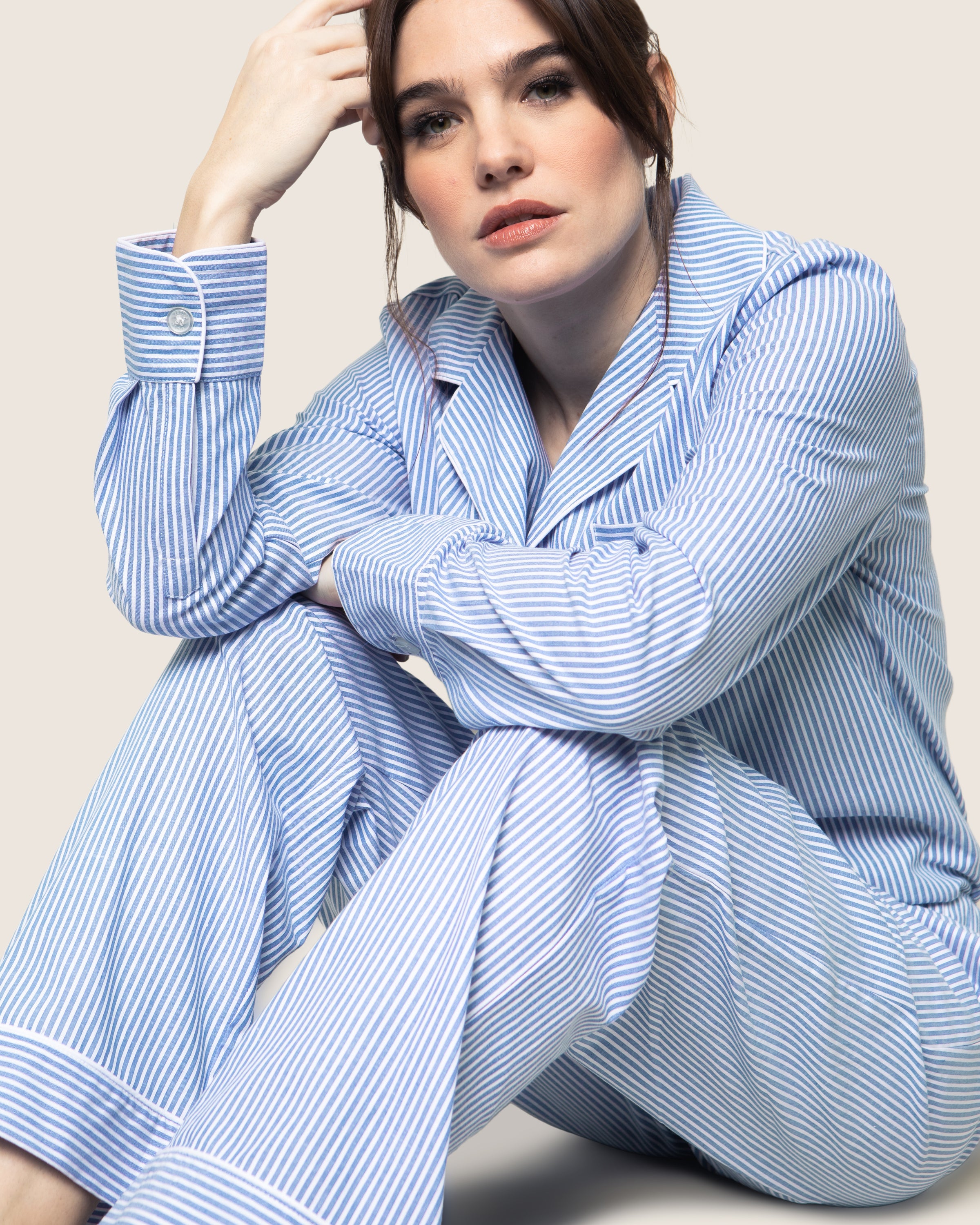 A woman in a Petite Plume Womens Twill Pajama Set in French Blue Seersucker sits against a neutral background, her elbow on her knee, gazing at the camera. The 100% cotton blue and white striped fabric adds texture to the scene.