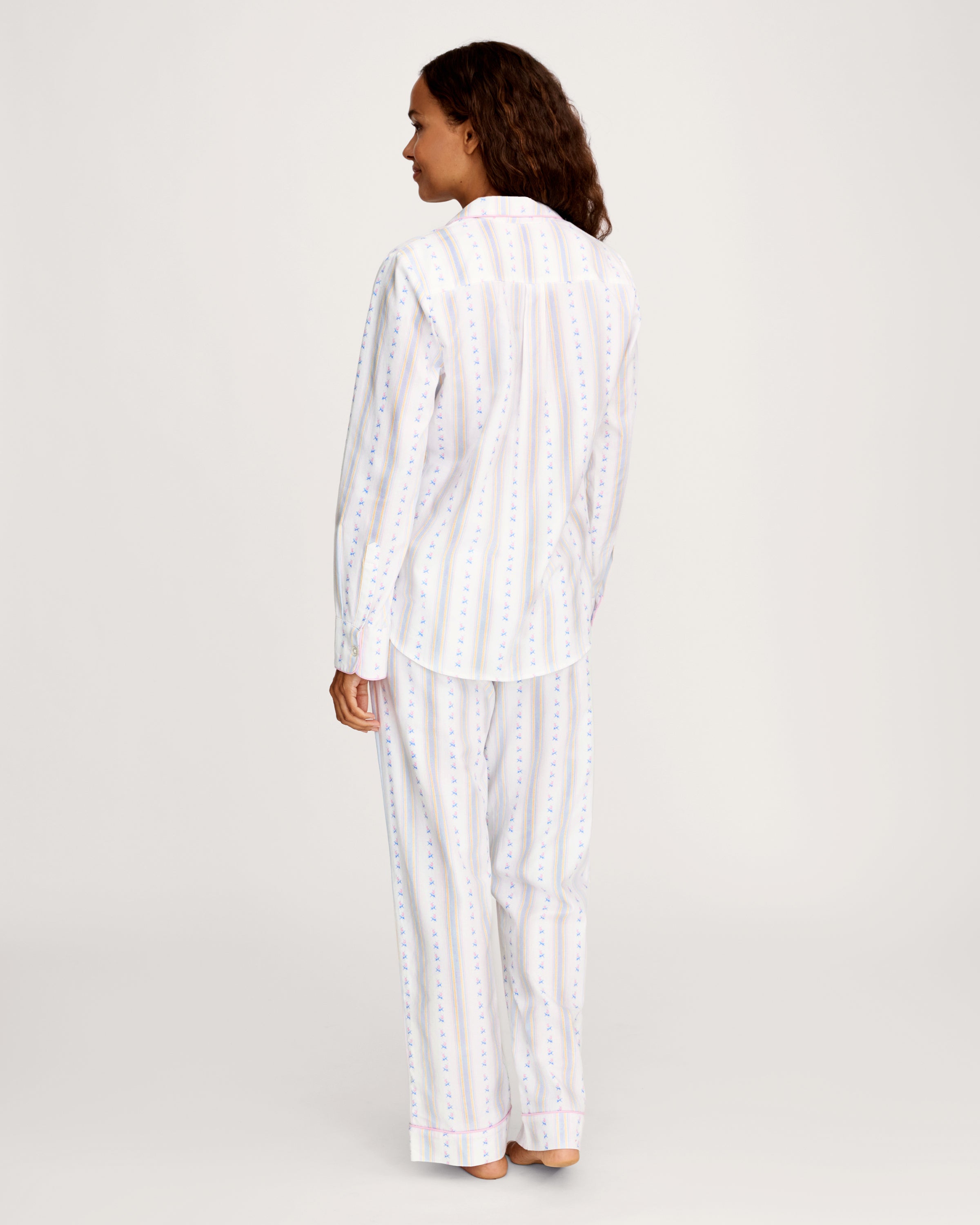 A woman with long curly hair stands barefoot against a plain light background, wearing the Women's Twill Pajama Set in Cottage Stripe—white pajama pants and a matching long-sleeve top with pastel vertical stripes and small dots.