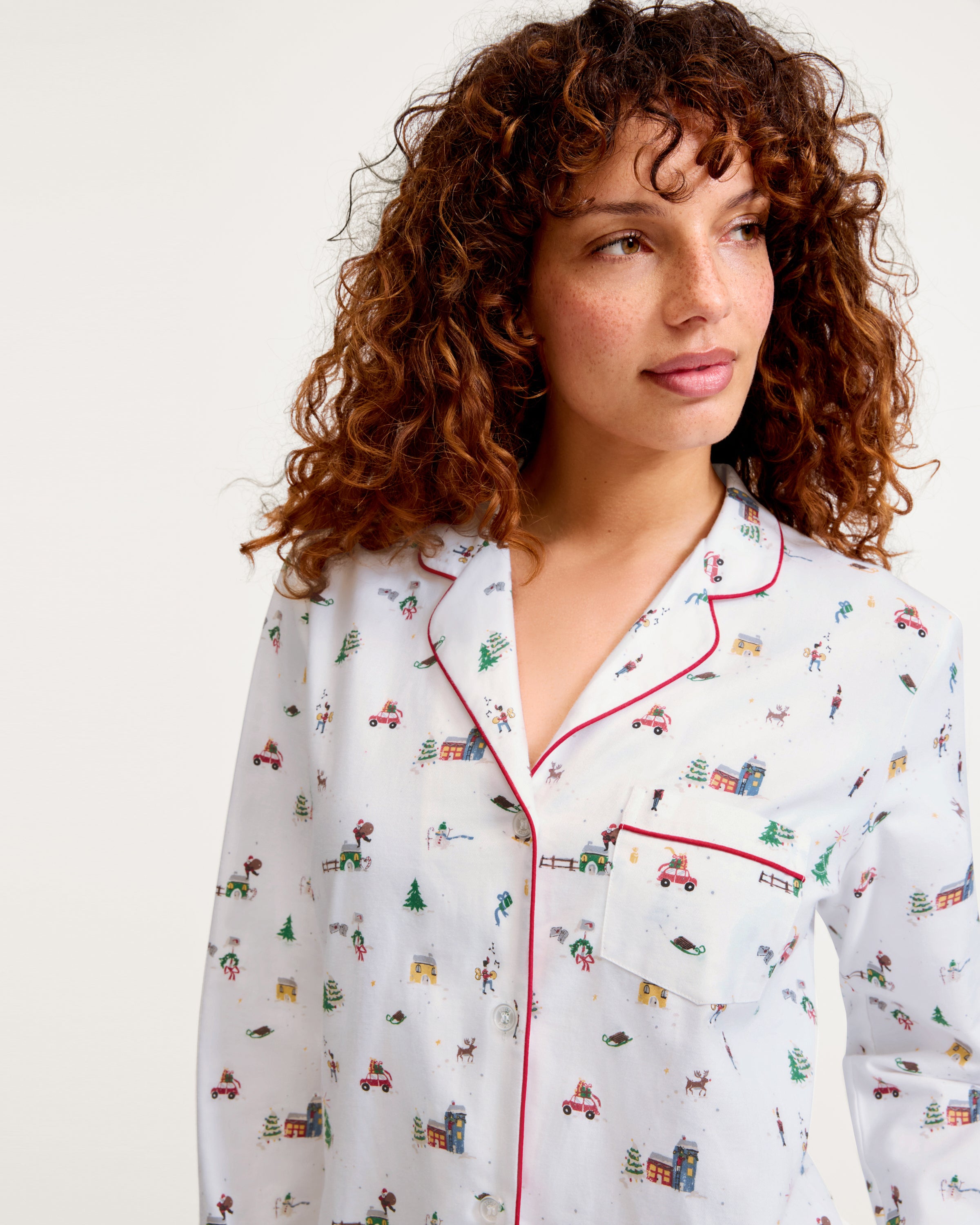 Women's Twill Pajama Set in Holiday Town: white pajama set with red piping, featuring festive prints of Christmas trees, cars, and skiers. Includes a long-sleeve button-up top with a pocket and matching drawstring pants.