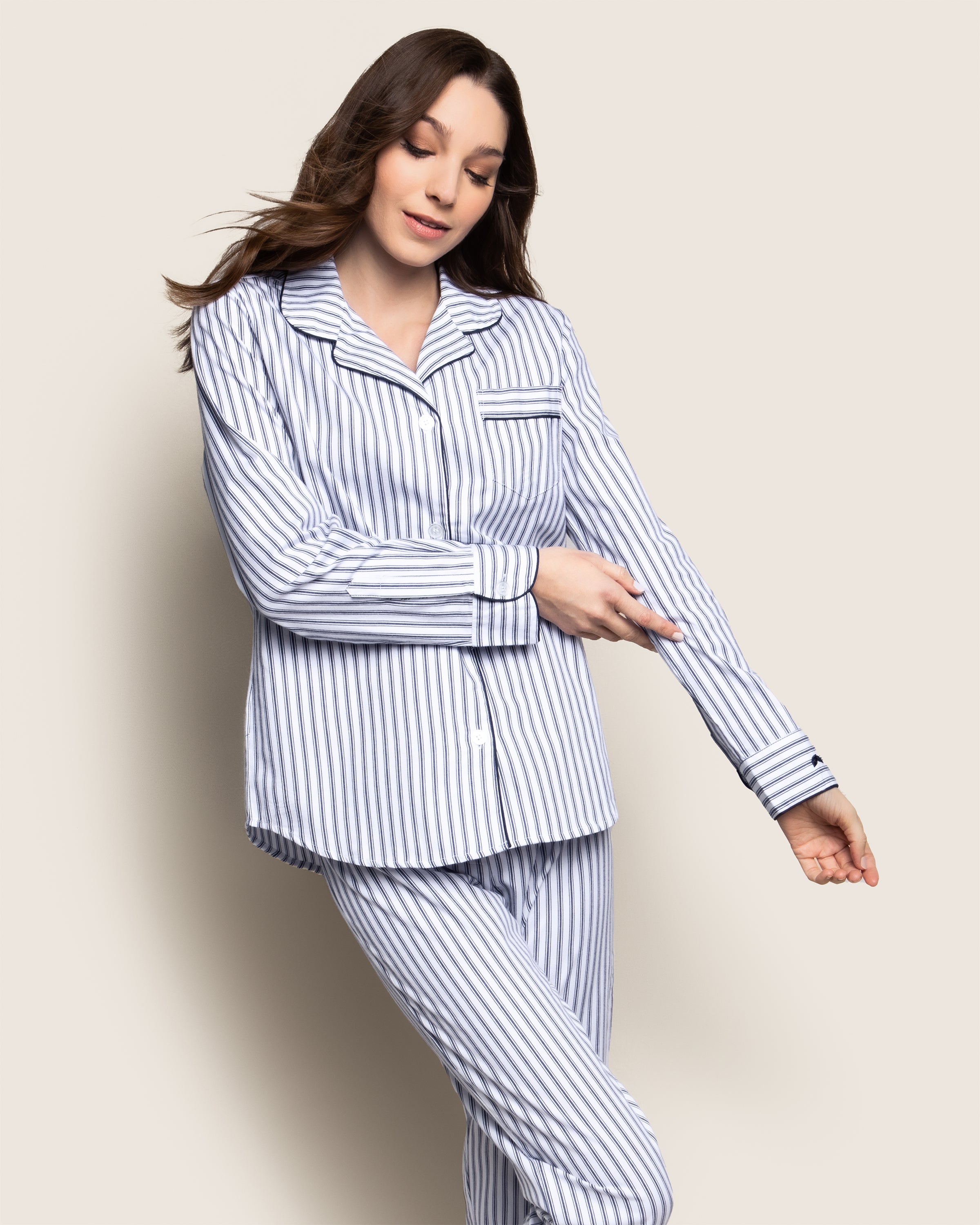A woman with long brown hair models the Petite Plume Womens Twill Pajama Set in Navy French Ticking. She points at her sleeve with a slight smile against a plain beige background.