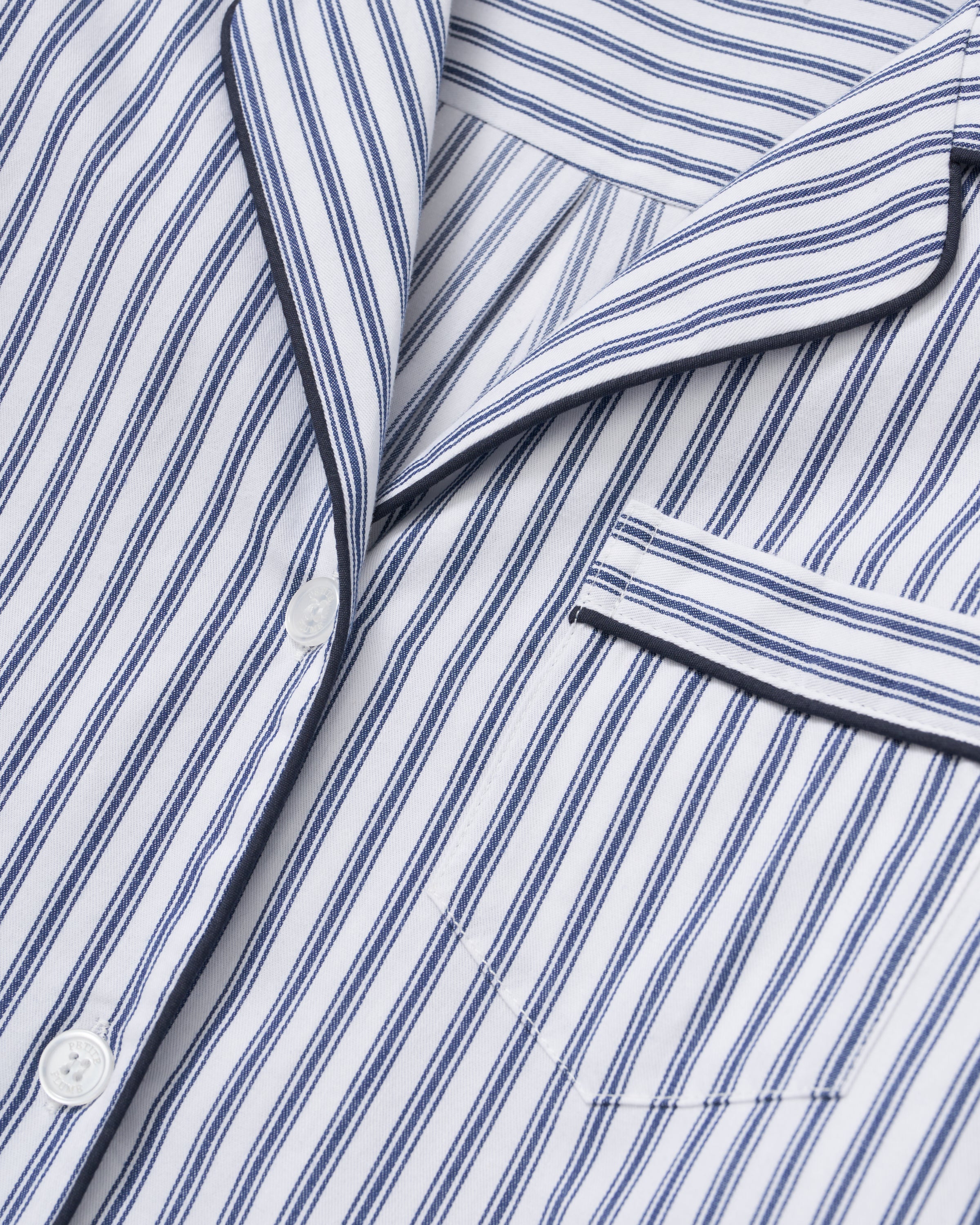 A model with long hair, wearing the Petite Plume Womens Twill Pajama Set in Navy French Ticking, poses against a plain background with hands in pockets. The cotton top features a collar and the pants are full-length, complementing her relaxed expression.