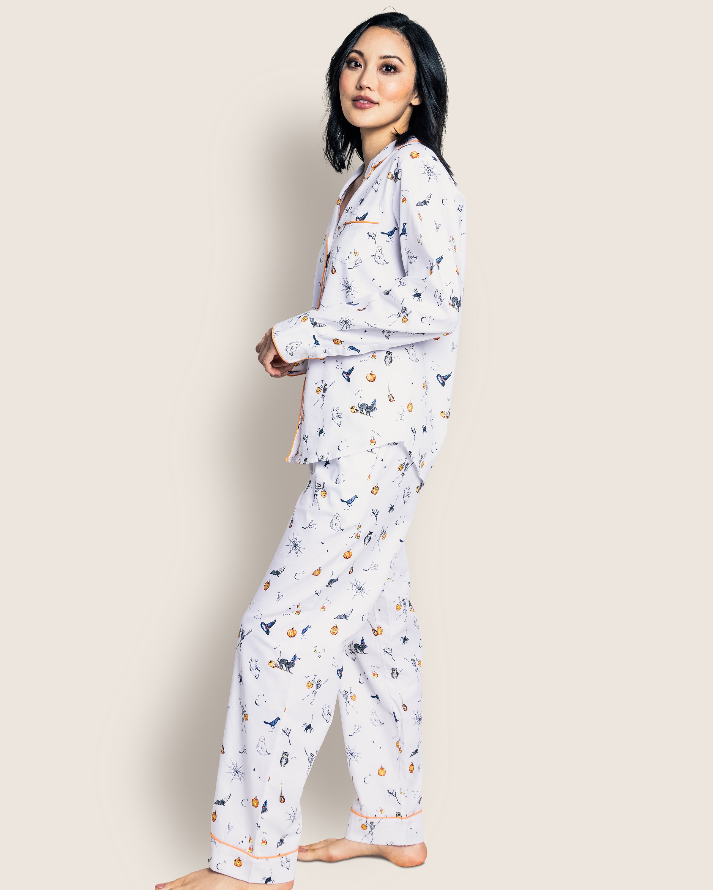 A person poses sideways against a plain background, smiling softly while wearing Petite Plumes Womens Twill Pajama Set in Trick or Treat. The cotton pajamas feature a matching white set with small Halloween prints, offering a relaxed fit with long sleeves and pants.