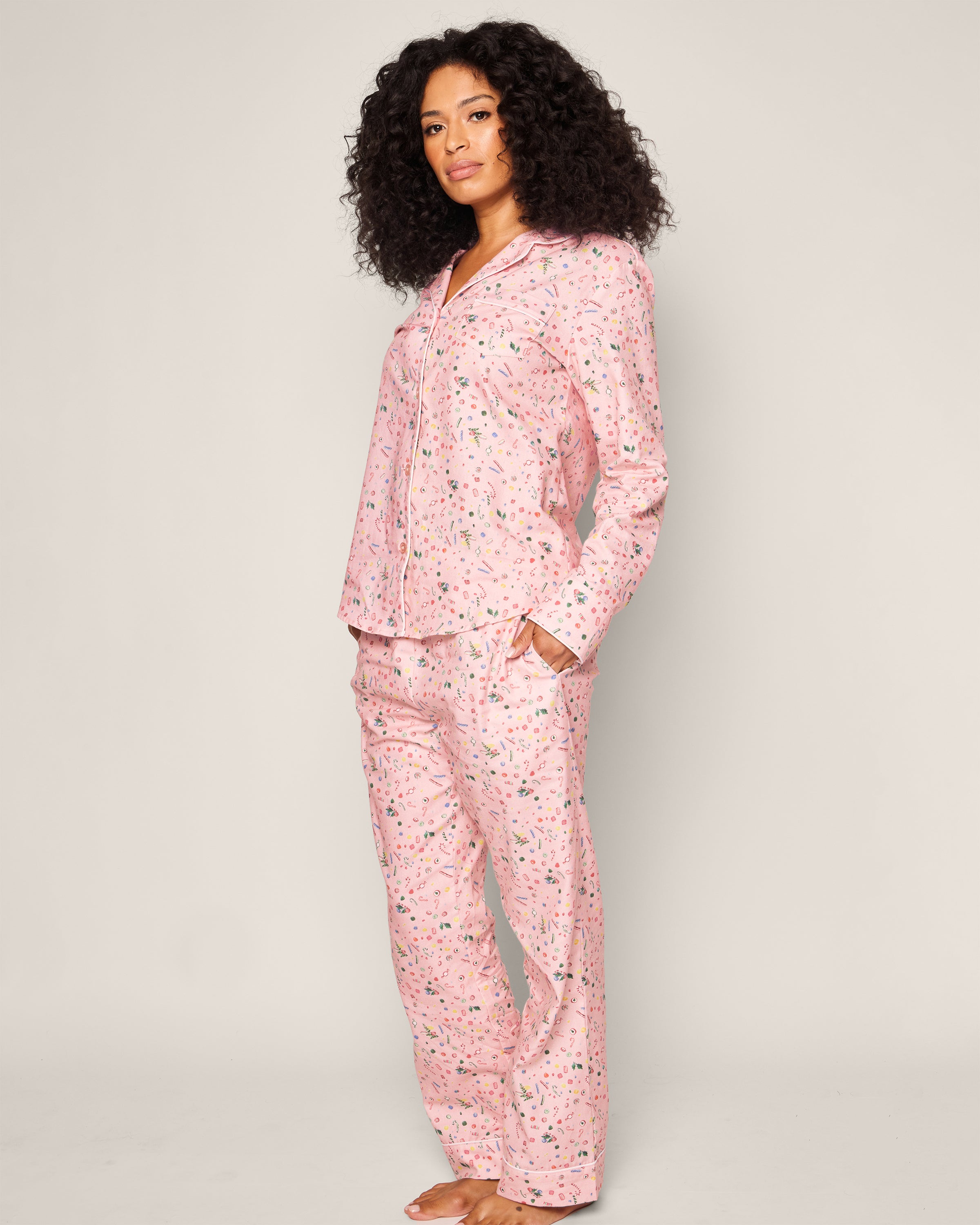 A curly-haired person stands facing forward in the Womens Twill Pajama Set in Vintage Sweets by Petite Plume. This cozy outfit features a pink cotton twill top with long sleeves and full-length pants, framed against a plain, light background enhancing its vintage charm.