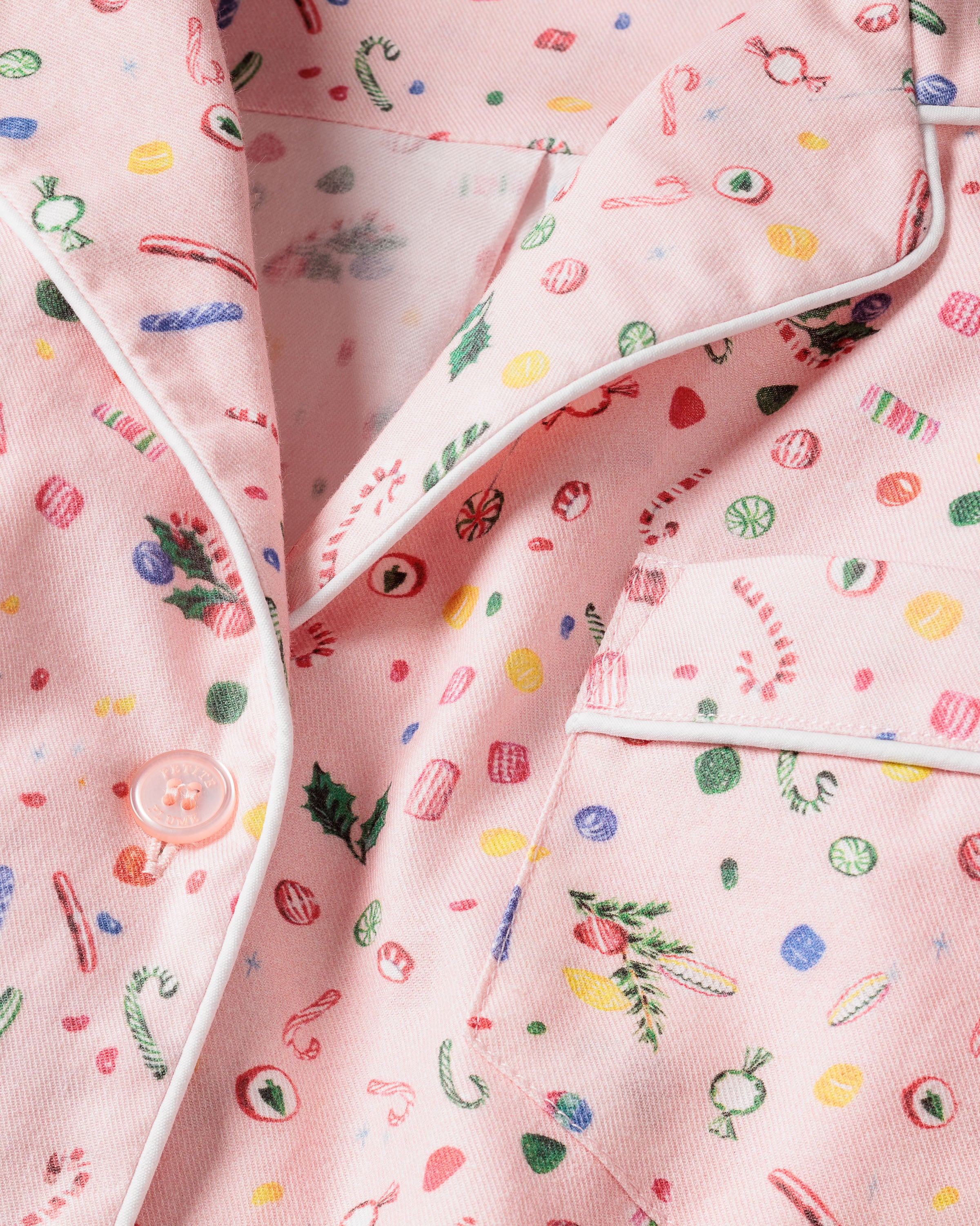 Close-up of pink fabric with candy canes, holly, and sweets creating a festive charm in this Womens Twill Pajama Set in Vintage Sweets by Petite Plume. The set includes white piping, a chest pocket, and a button for added detail.